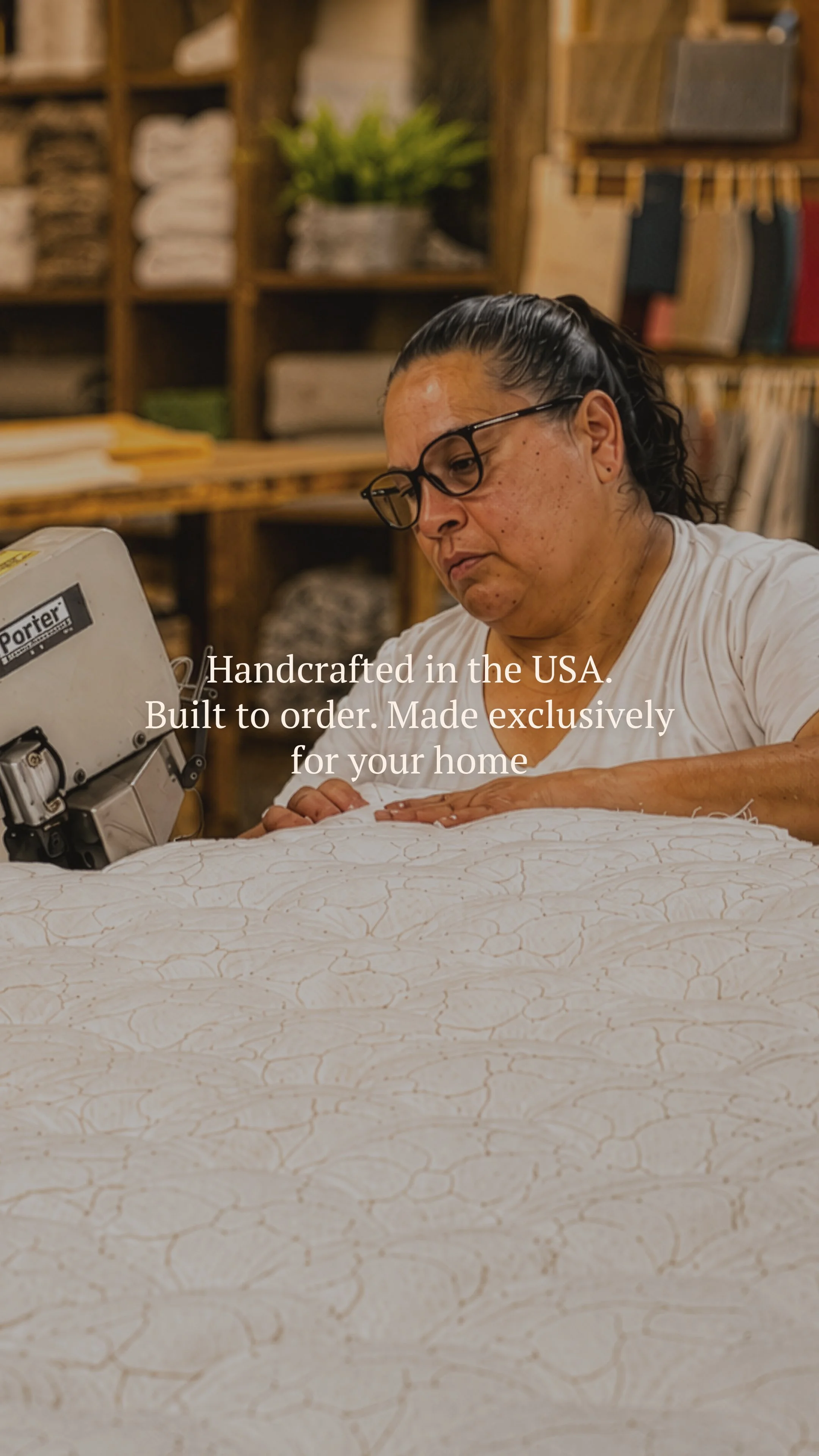 A woman with glasses and dark hair, working on a piece of fabric or material, in a workshop or studio with shelves of fabric and books in the background. The text overlay states: 'Handcrafted in the USA. Built to order. Made exclusively for your home