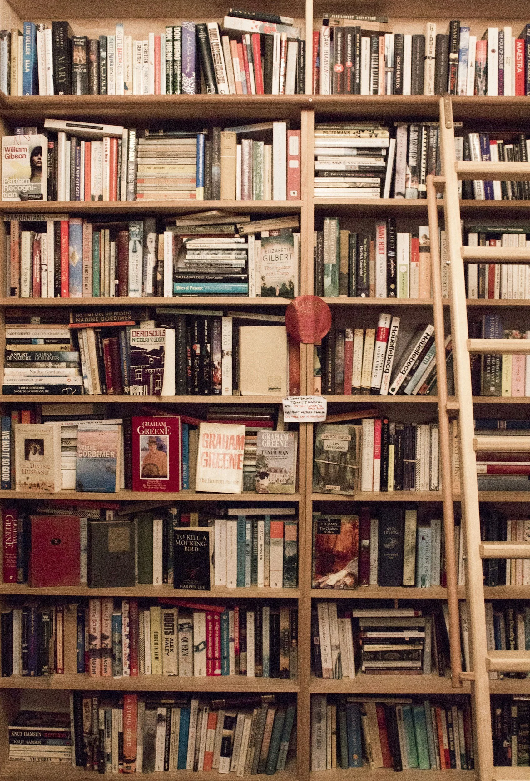A large wooden bookshelf filled with a variety of books in multiple rows and shelves, with a small wooden ladder on the right side.