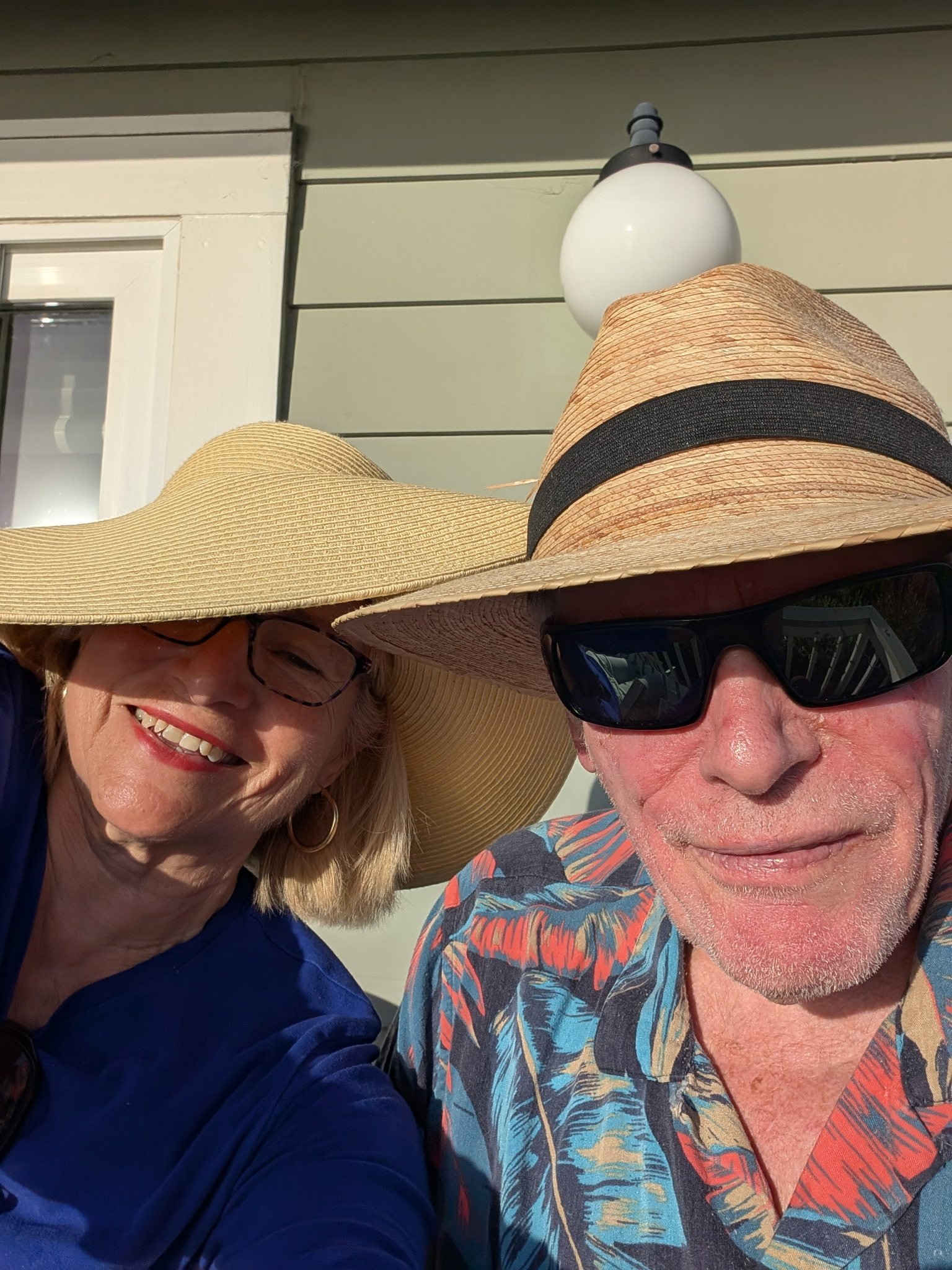 A smiling woman wearing a large straw sunhat and glasses, standing next to a man wearing a straw hat with a black band, sunglasses, and a colorful shirt with a tropical pattern, outdoors in front of a house with horizontal siding.