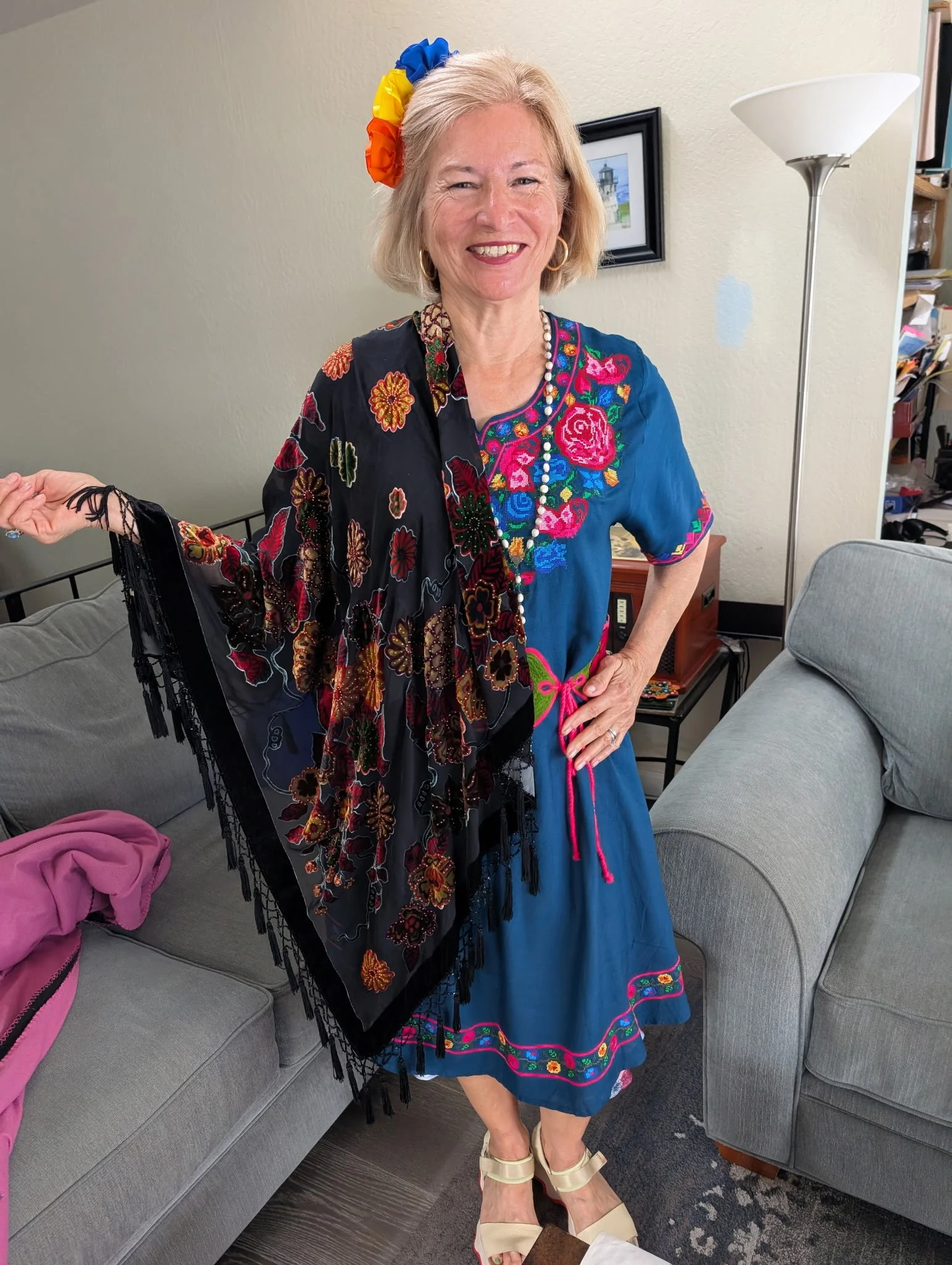 A smiling middle-aged woman with blonde hair wearing a colorful dress with floral embroidery and a bead necklace. She has a rainbow-colored flower hair accessory and is posing indoors with a black and white picture on the wall, a white floor lamp, and a grey couch with a pink cloth on it.