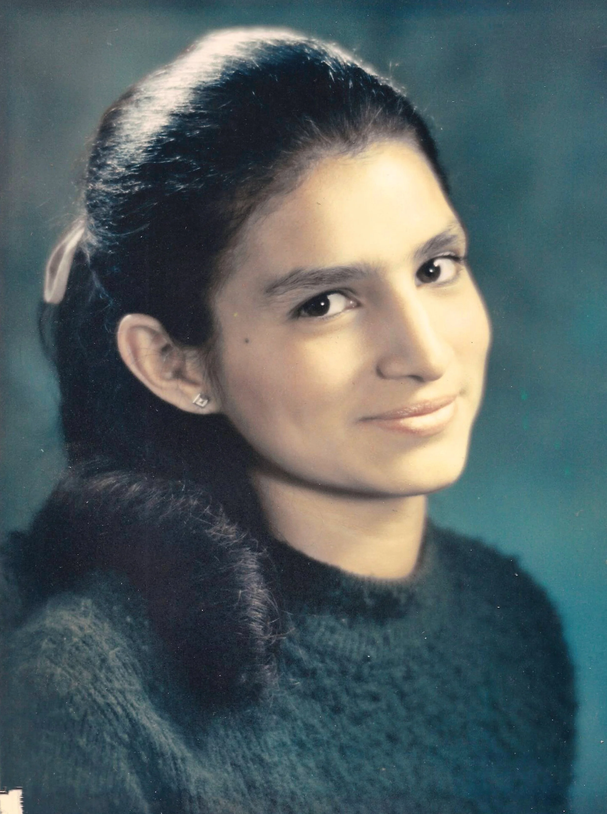 A portrait of a woman with dark, wavy hair styled back, smiling softly with a dark background.