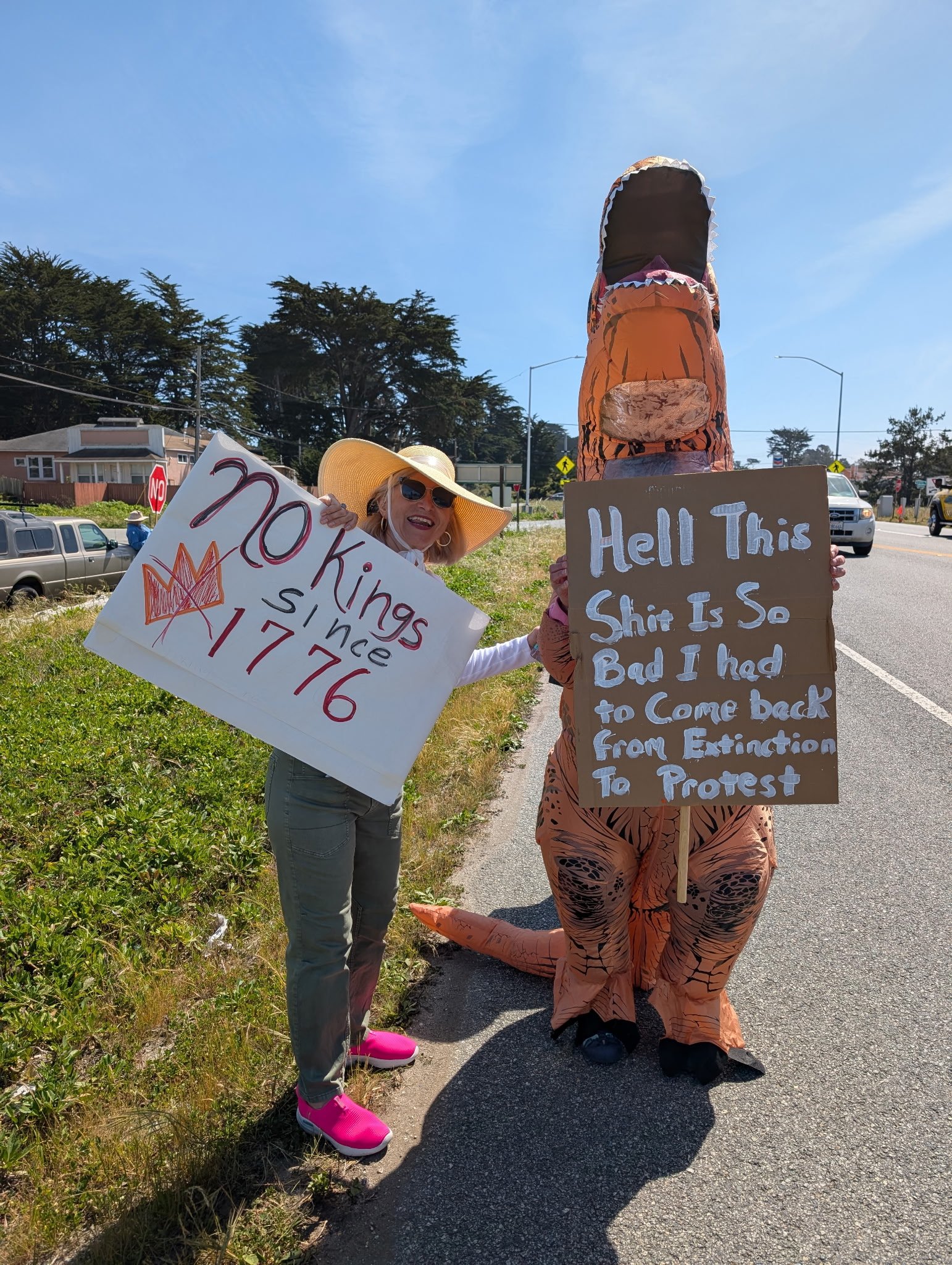 People protesting on the street, with one person dressed in a dinosaur costume and holding a sign that reads, 'Hell This Shit Is So Bad I Had to Come Back From Extinction to Protest'. Another person is holding a sign that says, 'No Kings Since 1776'.