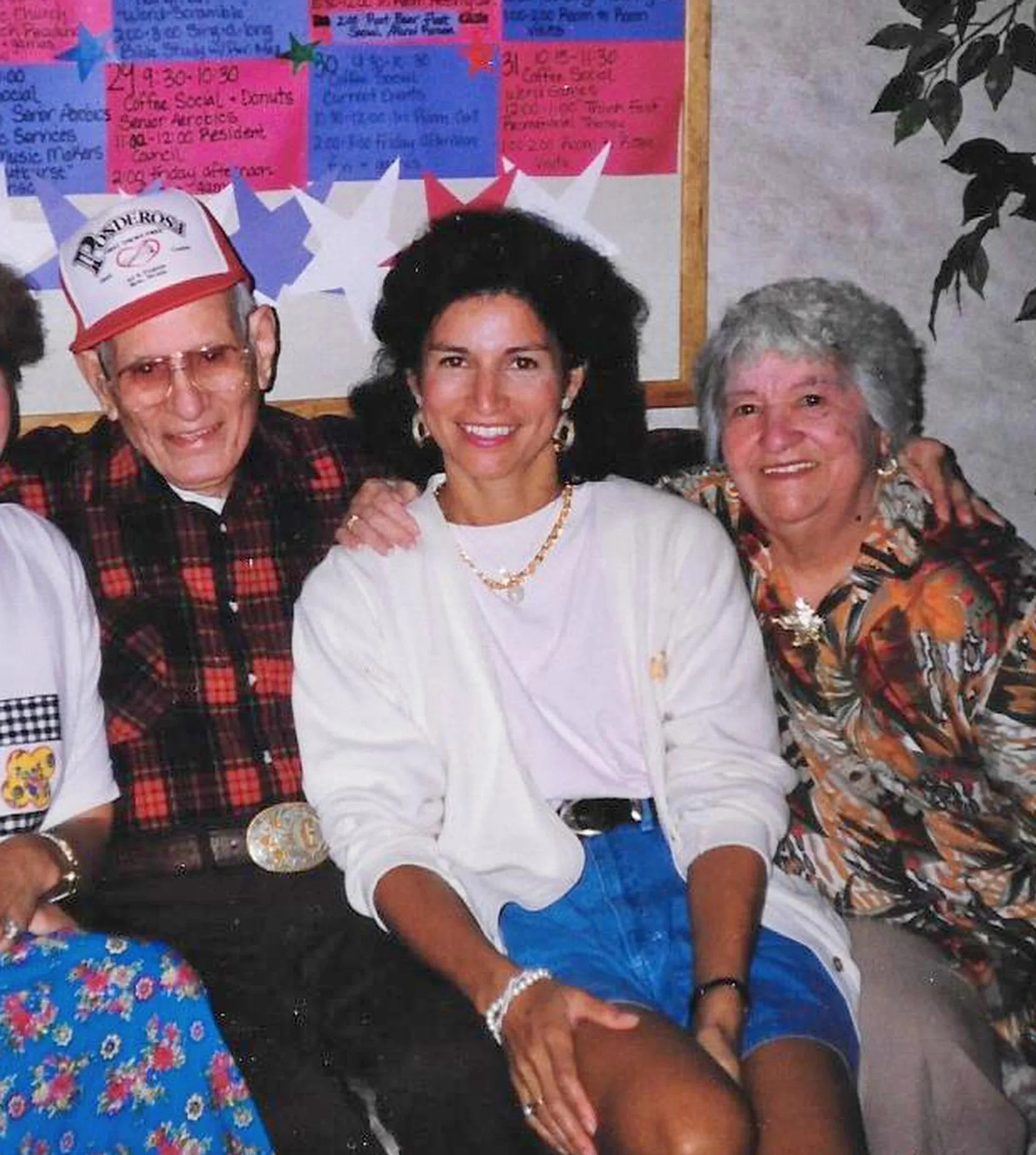 Three people smiling for a photo, with two women on the right and an elderly man on the left, dressed casually. The man is wearing a red and white hat and glasses, while the women are wearing jewelry. The background features colorful posters and a gray wall, with some green leaves visible.