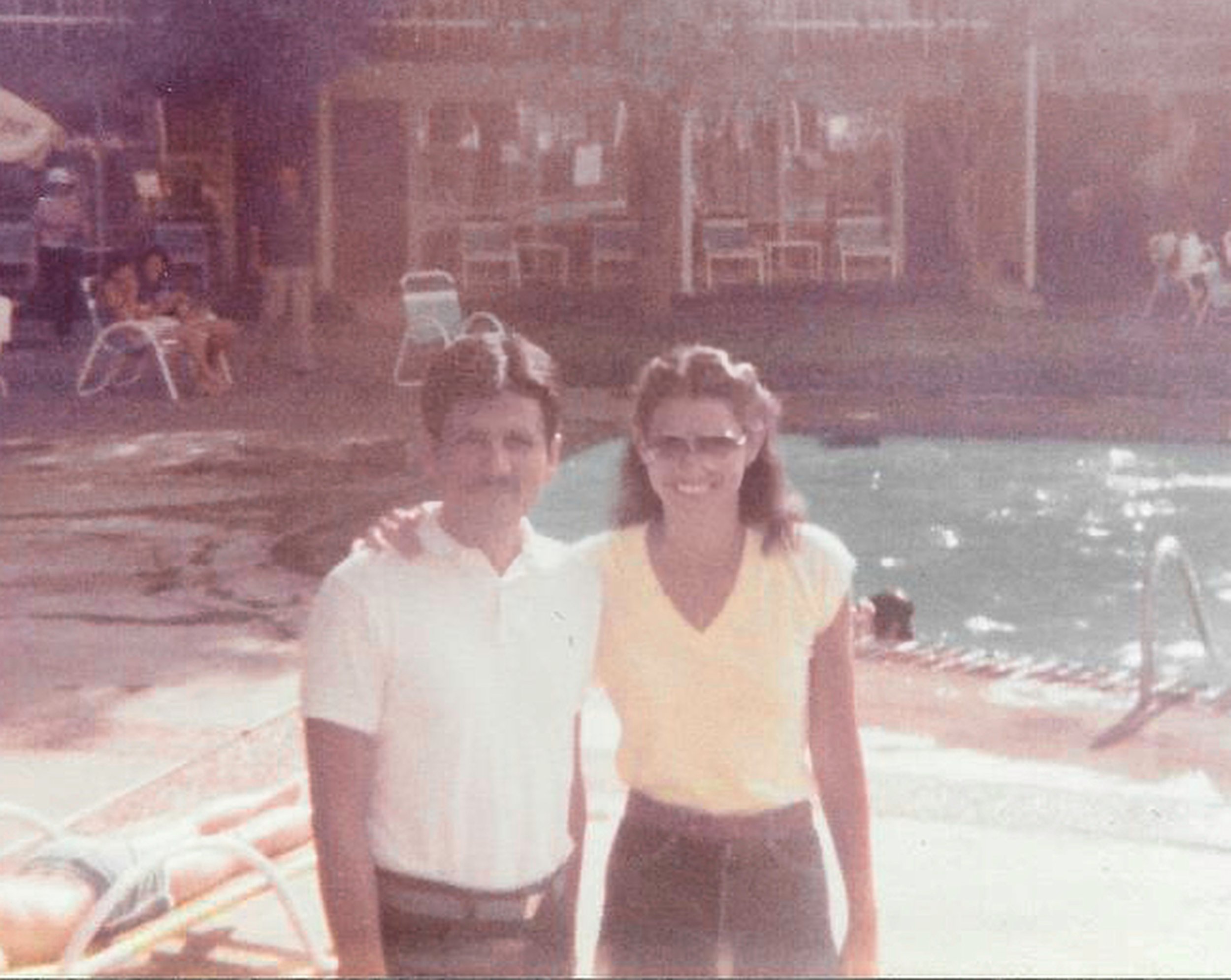 A vintage photograph of two young people smiling and standing close to each other on a beach, with a house and chairs in the background.