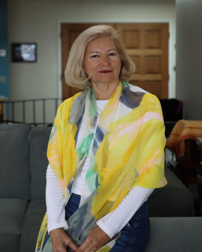 An older woman with blonde hair standing in a living room, wearing a multicolored scarf and white long-sleeve shirt.