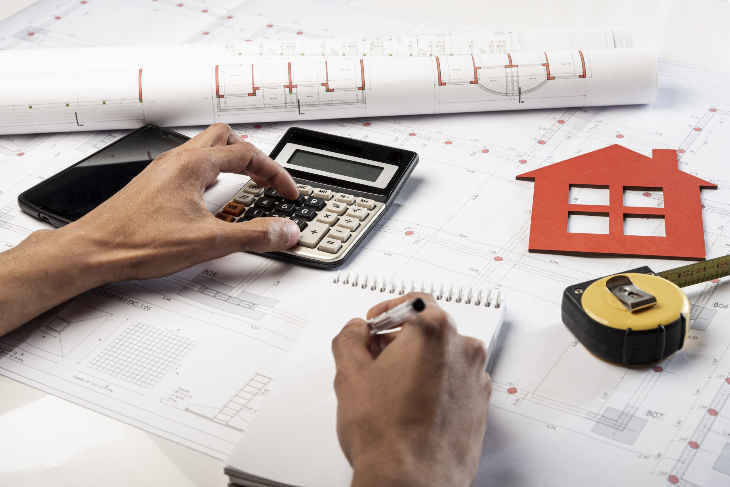 Architectural blueprints and drawings on a table with a hand using a calculator and another hand writing, along with a small house model and measuring tape.