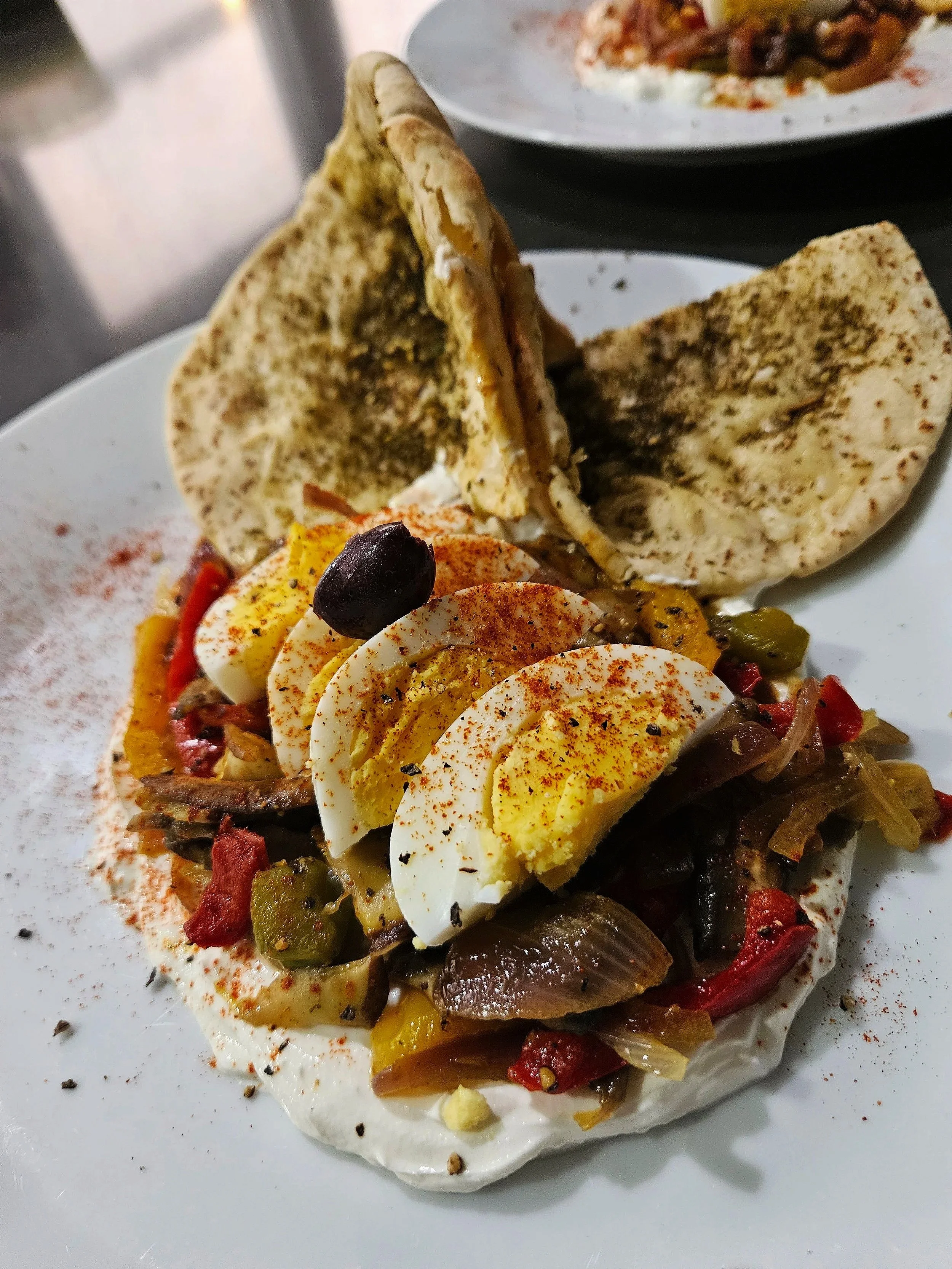 Open-faced Srian breakfast. Served on a bed of Labneh, topped with  with sliced hard-boiled eggs, red and green peppers, olives, and sprinkled with paprika. Served on the side with a Pita bread, Zatar bite.