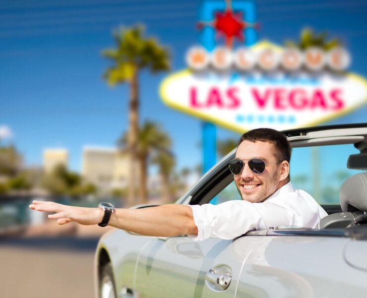 A man with sunglasses smiling and waving from a convertible car in front of the Las Vegas sign with palm trees and a blue sky in the background.