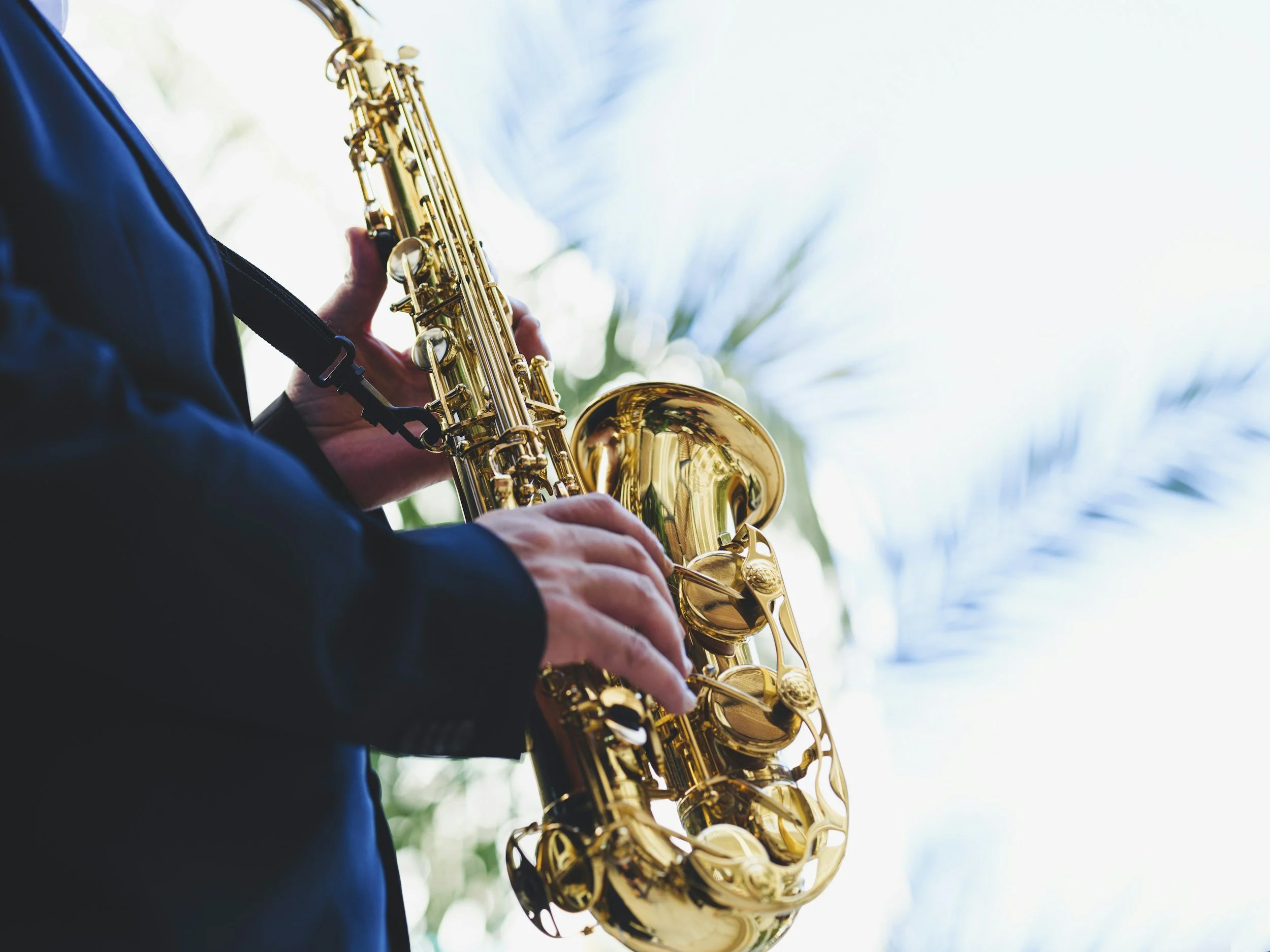 A person playing a shiny gold saxophone outdoors, wearing a dark jacket, with a blurred background of greenery and sky.