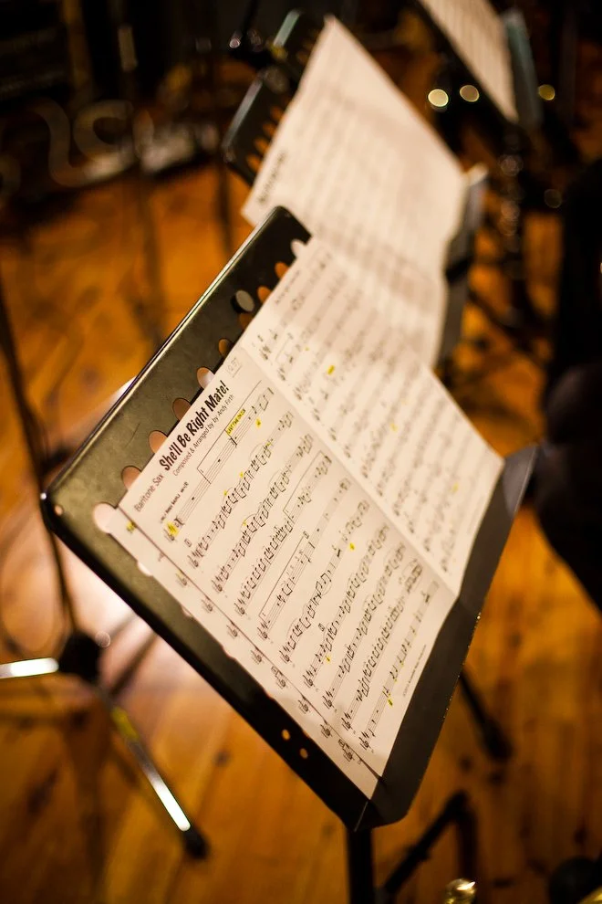Music stand holding sheet music in a dimly lit room with wooden floors and chairs.