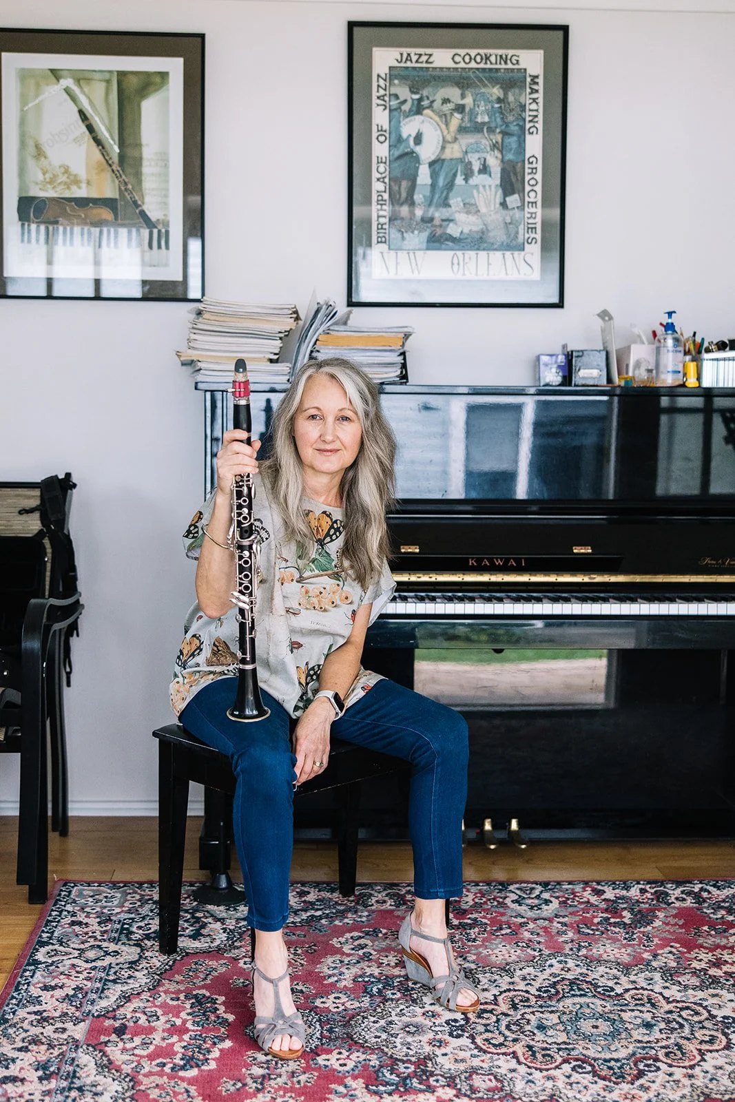 A woman sitting on a black chair holding a clarinet in a room with a black grand piano, framed jazz-themed posters on the wall, a basket of pens and bottles on the piano, and papers stacked in the background.