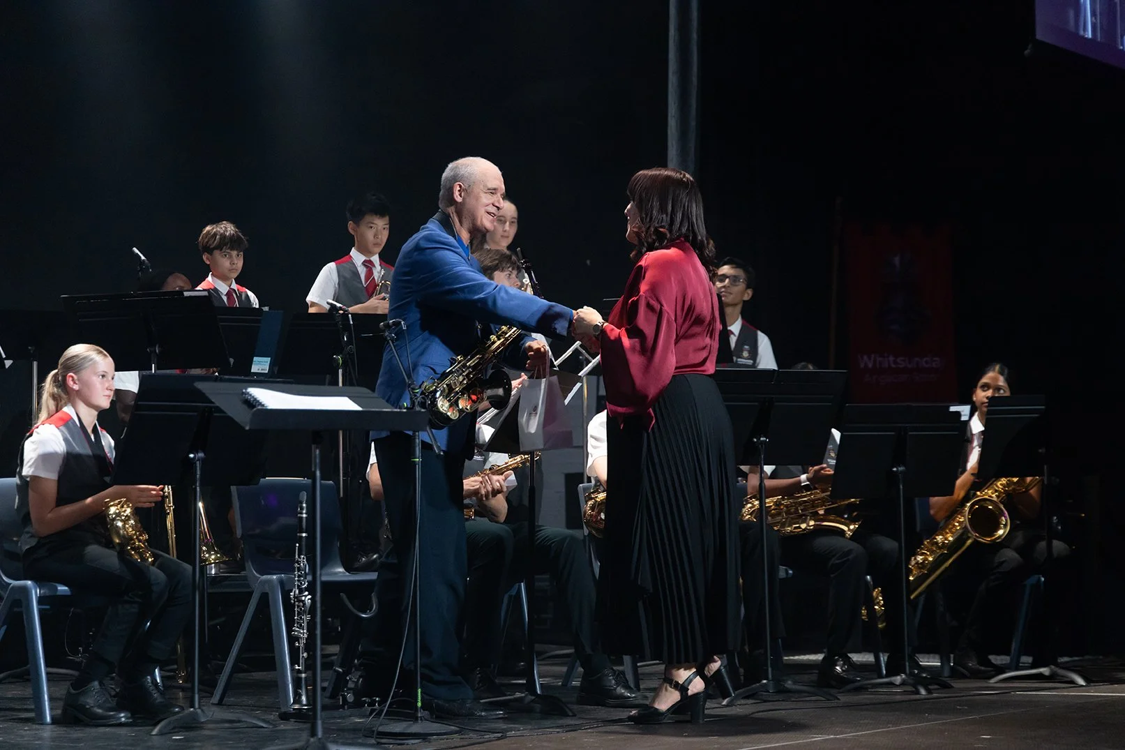 Andy Firth in a blue suit handing an award to a woman in a red blouse on stage with a school band and students in uniform in the background.