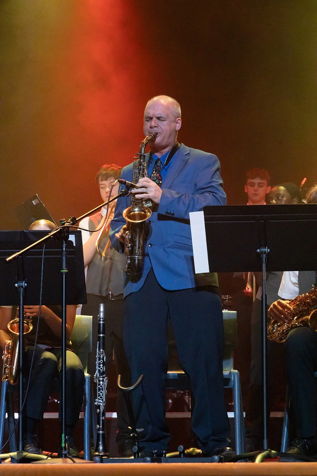 Andy Firth in a blue suit jacket playing the saxophone during a concert with a group of young musicians on stage, illuminated by warm stage lighting.