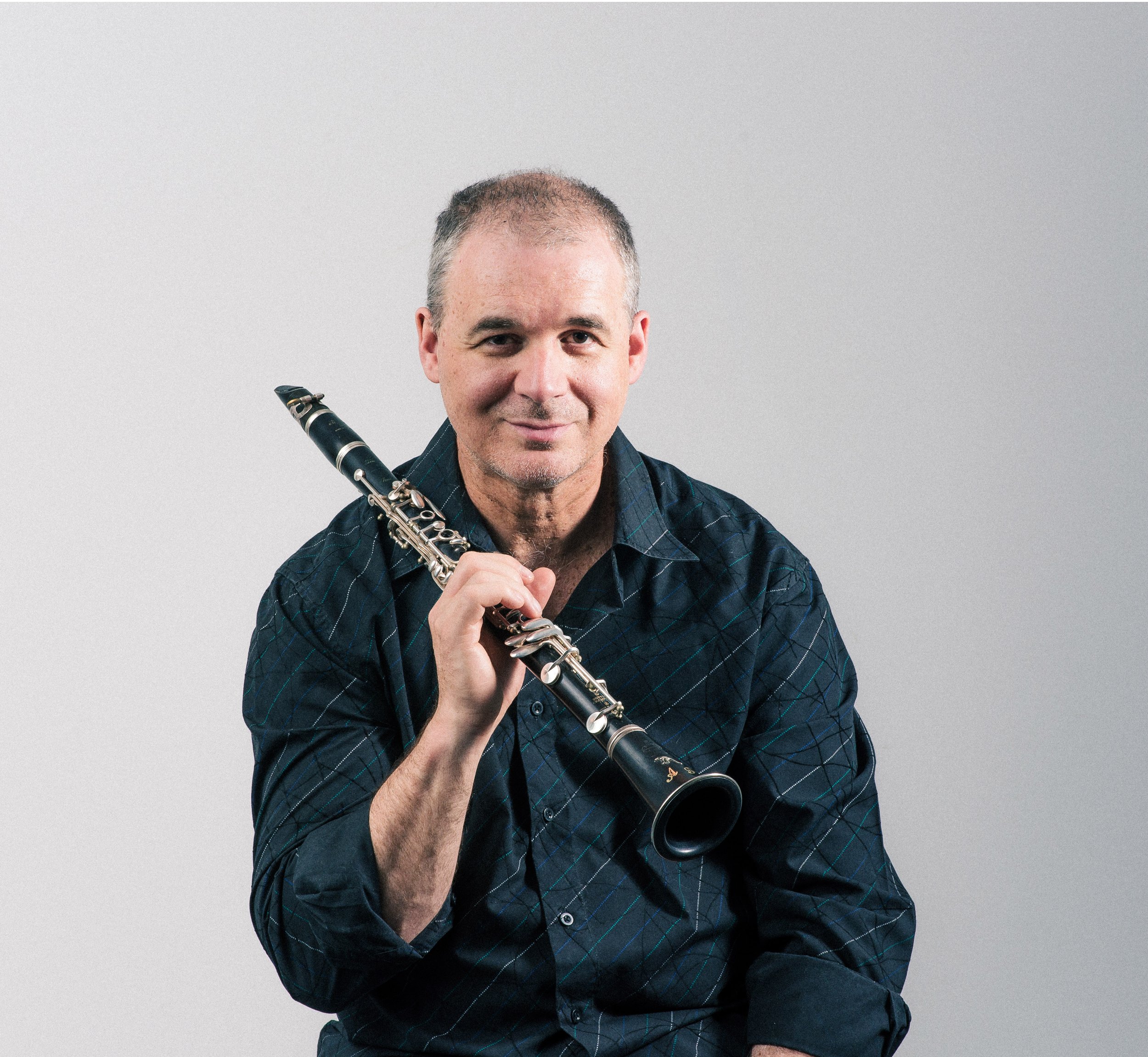 A middle-aged man with short gray hair and a goatee, smiling gently while holding a clarinet, standing against a plain light gray background.