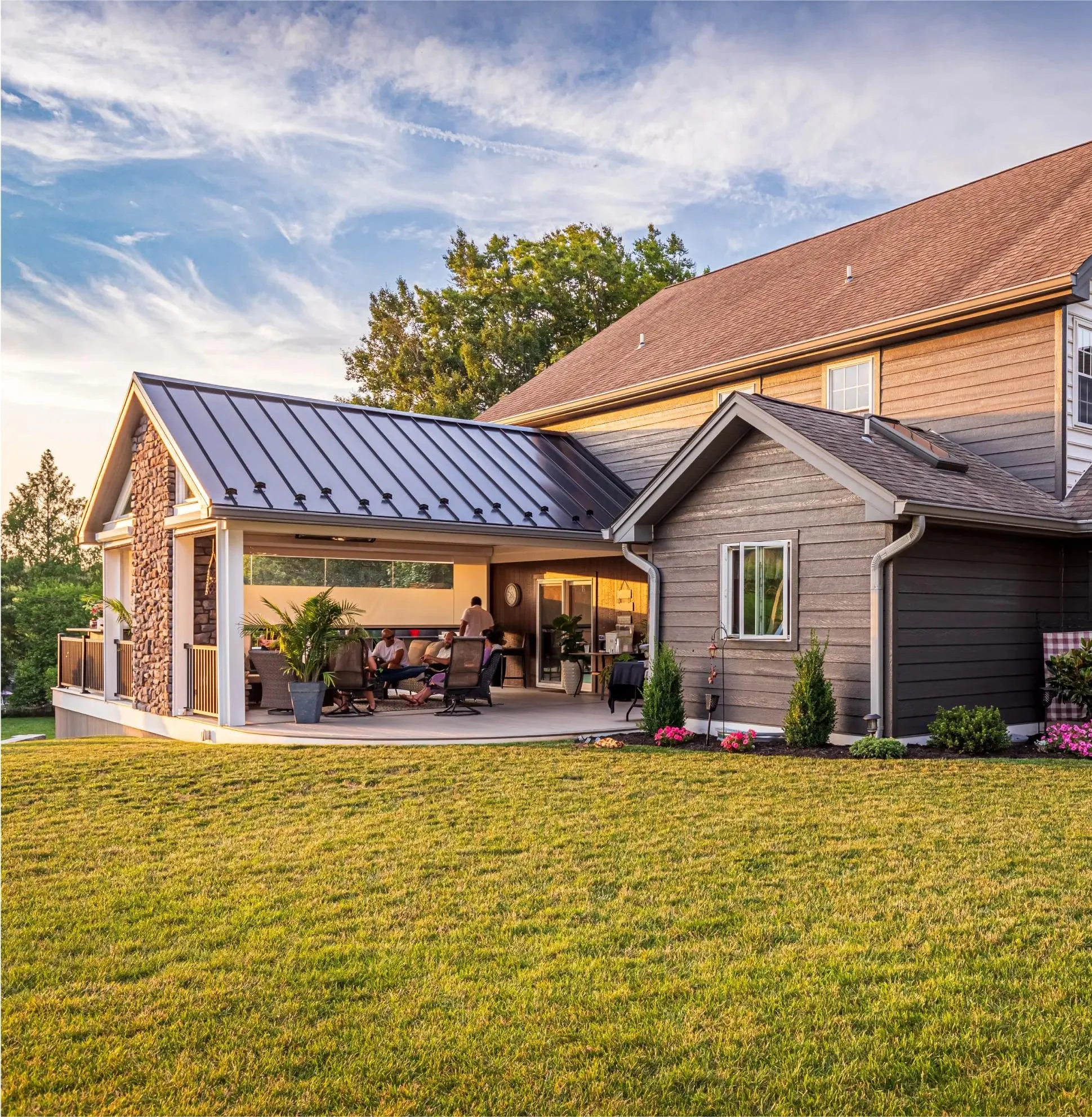 A house with a backyard patio area, multiple seating chairs, and potted plants, with a well-maintained grassy lawn, under a partly cloudy sky in the evening.