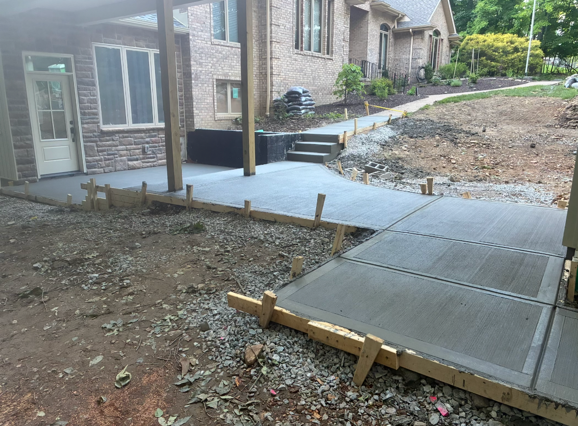 Residential construction site with newly poured concrete sidewalk and stairs, wooden forms still in place, in front of brick houses, with landscaping and dirt areas surrounding the walkways.