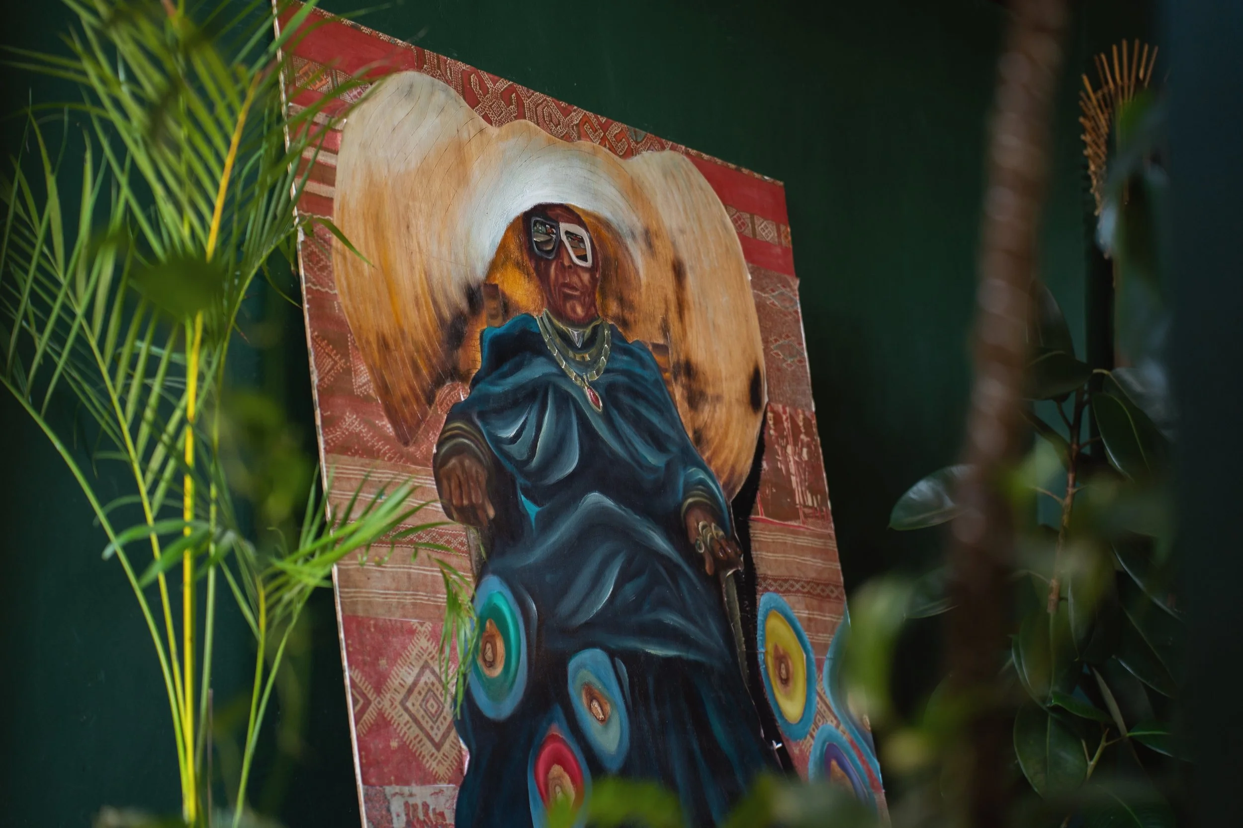 Colorful artwork of a person wearing glasses, seated with a vibrant background, partially obscured by green foliage.