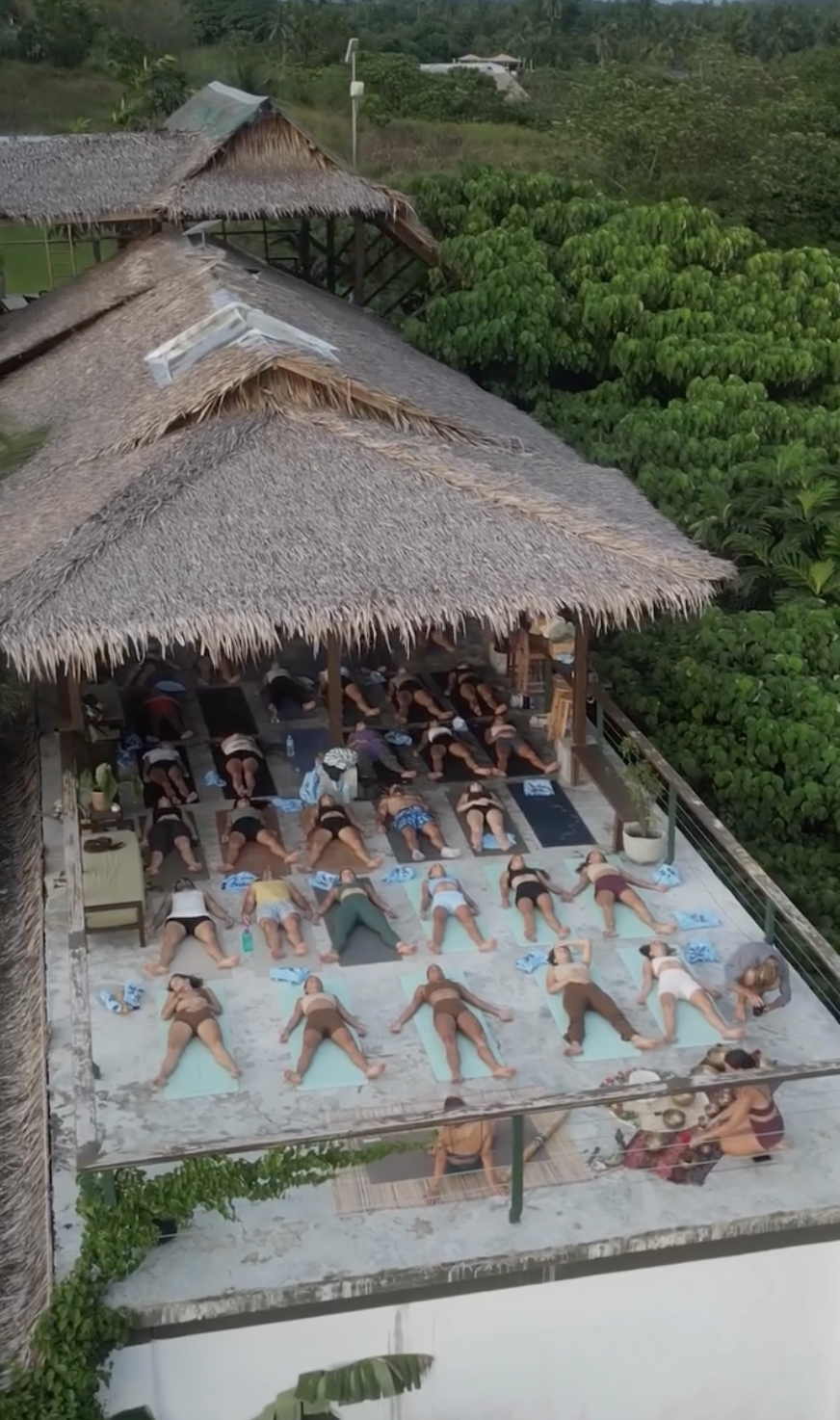 Group of people practicing yoga on mats under a thatched roof in an outdoor setting surrounded by lush green trees.