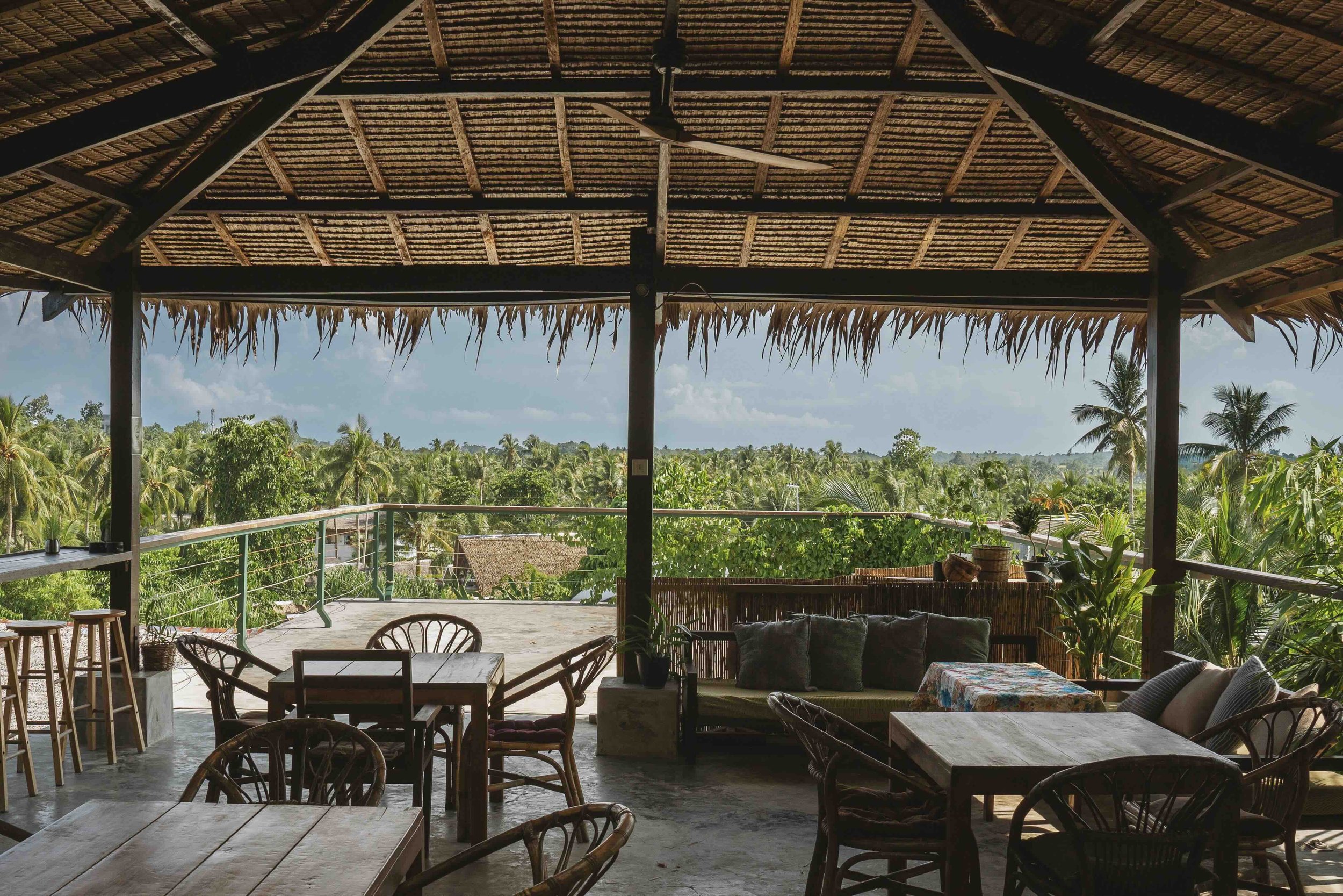Open-air cafe or restaurant with tropical plants, thatched roof, and outdoor furniture overlooking lush greenery and palm trees.