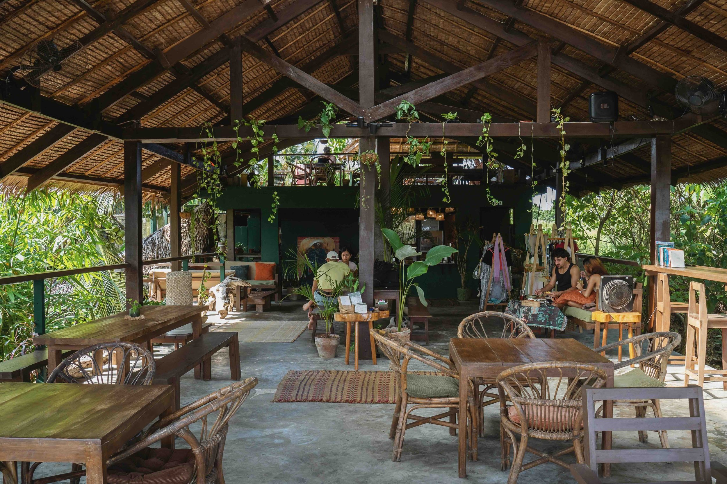 Open-air, rustic cafe or restaurant with wooden furniture, plants, and tropical decor, with people sitting and chatting.