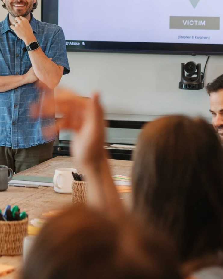 People in a meeting room with a presentation slide on the screen that says "Victim" and a person standing near the screen with crossed arms.