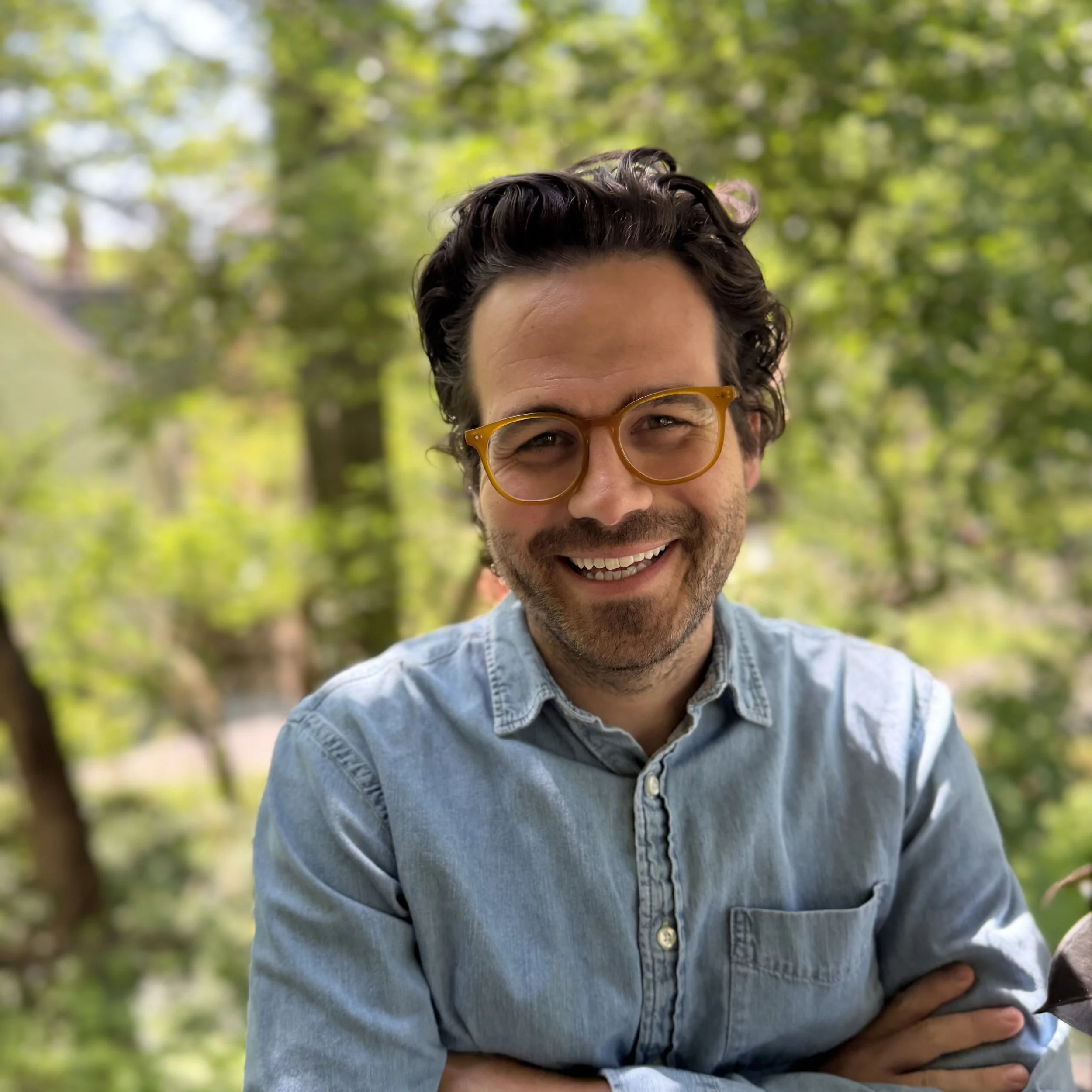 Close-up of a smiling man with curly dark hair and glasses, wearing a light blue denim shirt, standing outdoors with green trees in the background.