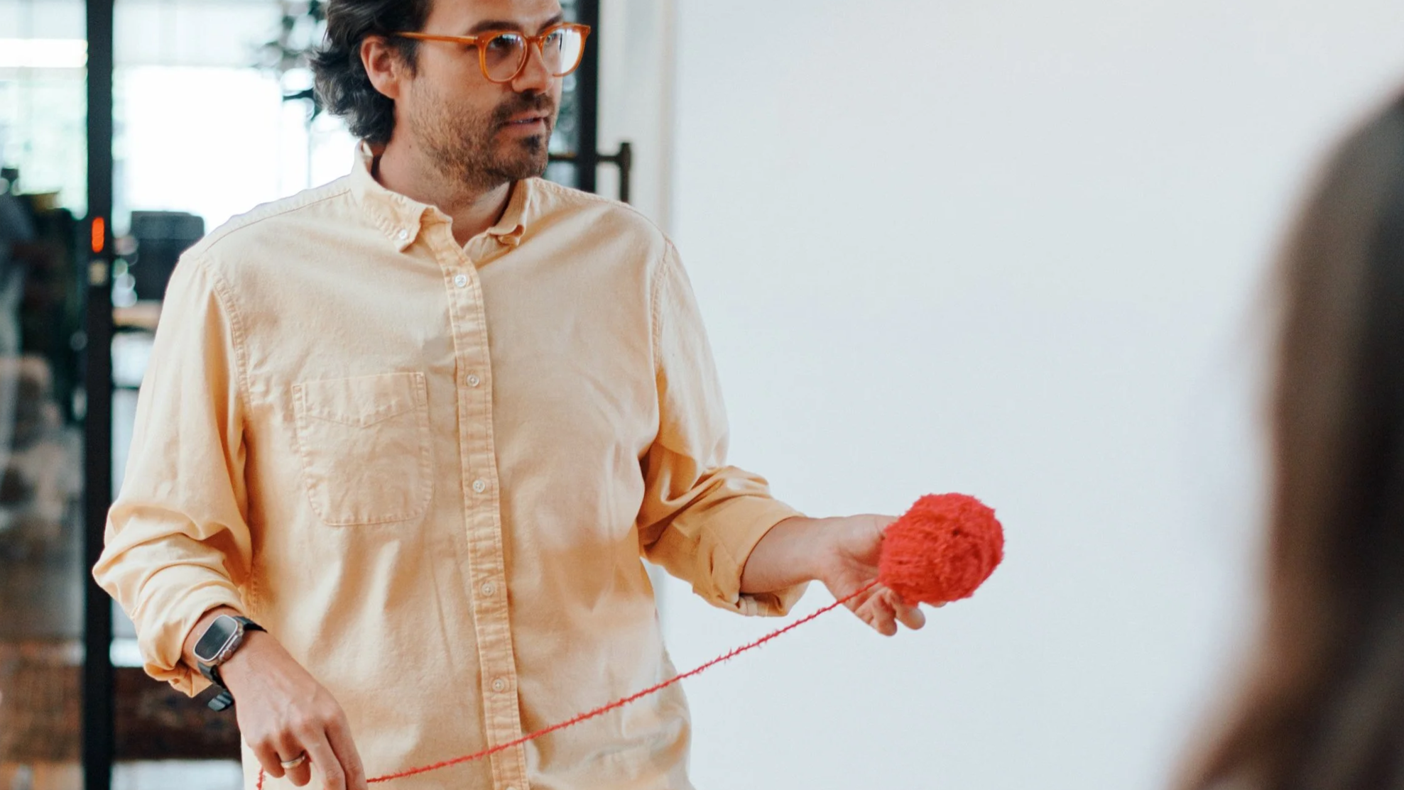 A man wearing an yellow shirt with glasses holding a orange ball of yarn in an indoor setting.