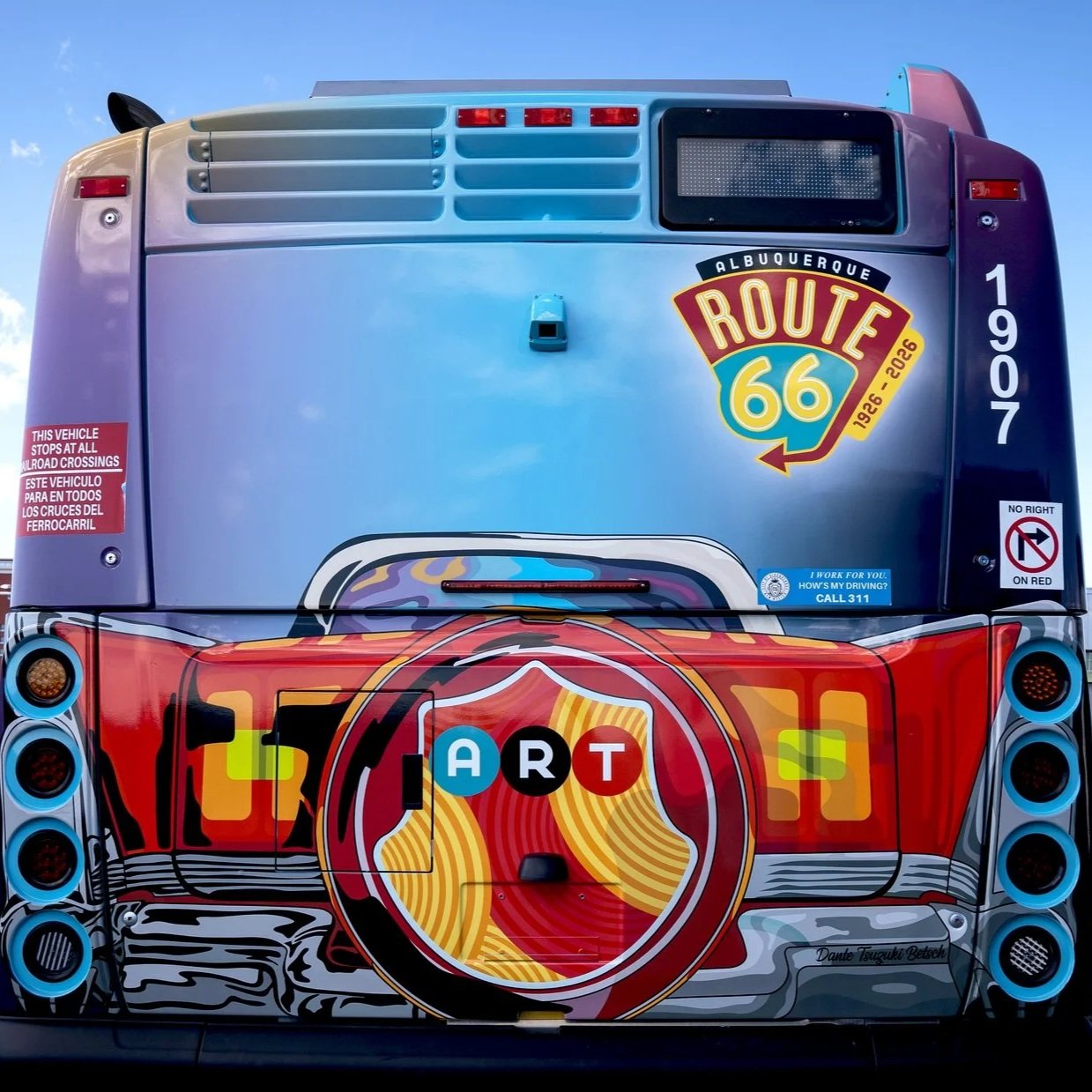 The back of a colorful bus featuring art inspired by Route 66, with the word ART replaced by colorful balls, and various signs including one for railroad crossings and a phone number for driver safety.
