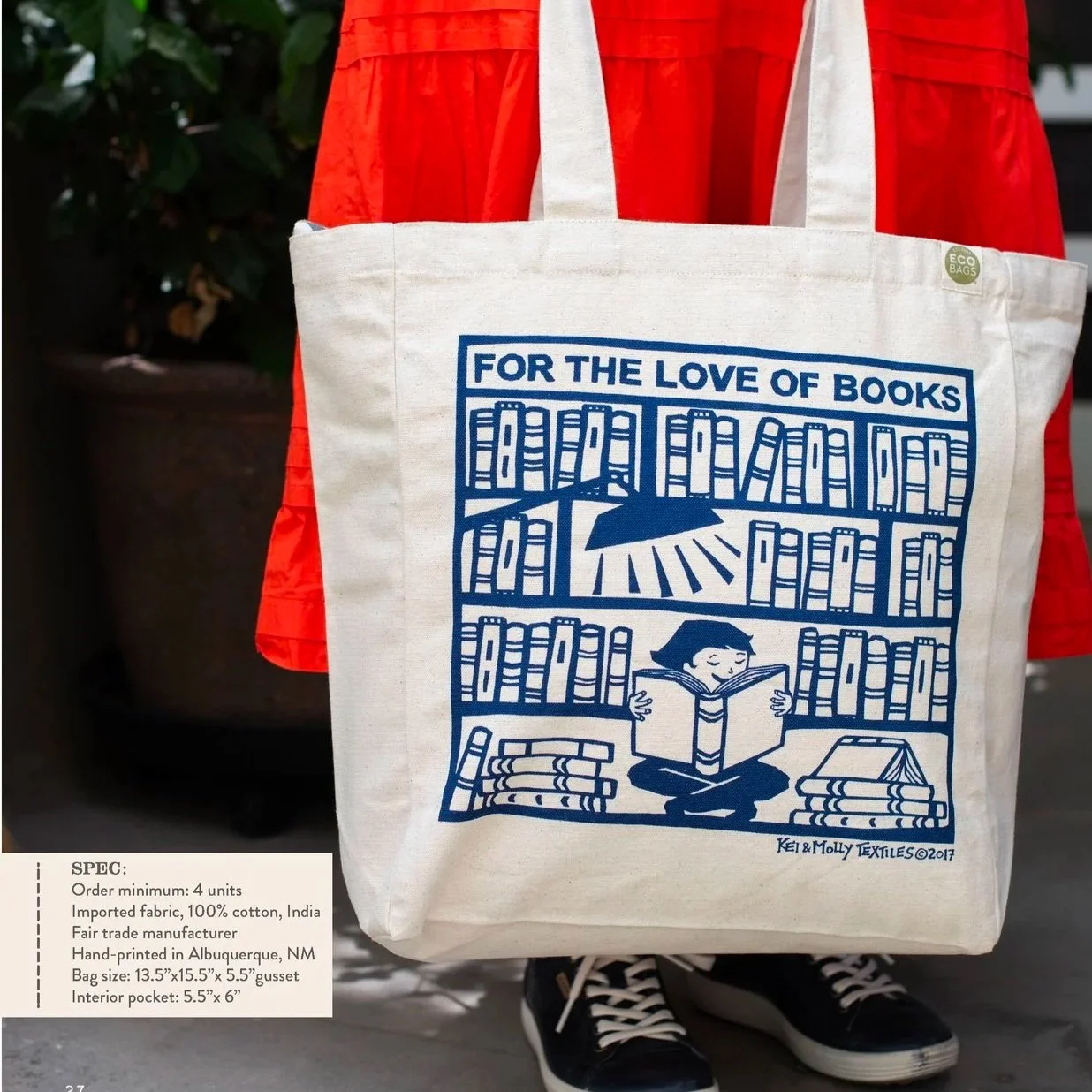 A tote bag with a blue print design reading 'For the love of books', featuring a child reading amidst bookshelves with an umbrella and rays of light, against a background of a person in a red skirt and black shoes.