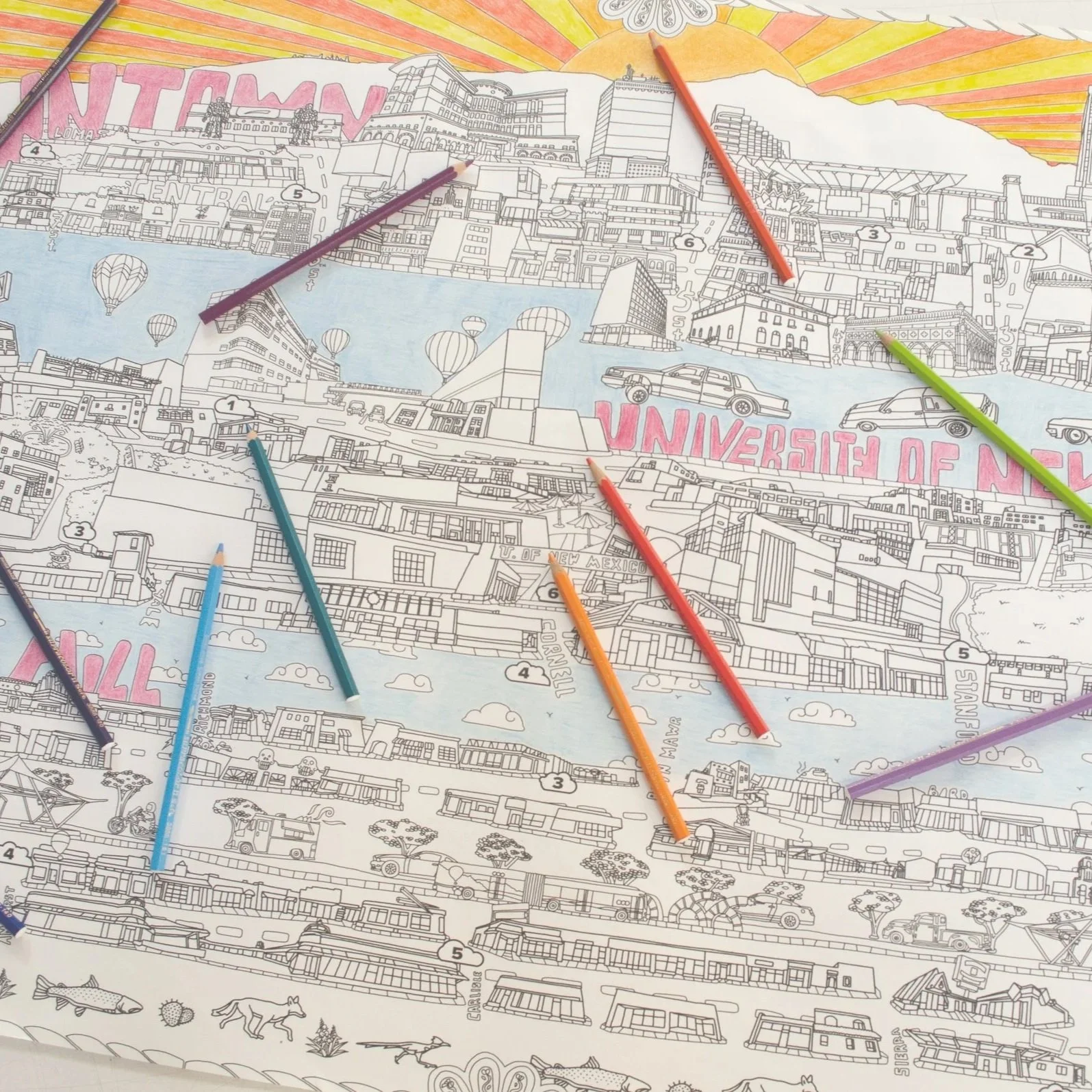 Coloring map of a cityscape with various buildings, vehicles, hot air balloons, and the text 'UNIVERSITE DE NE' visible, with colored pencils placed on the map.