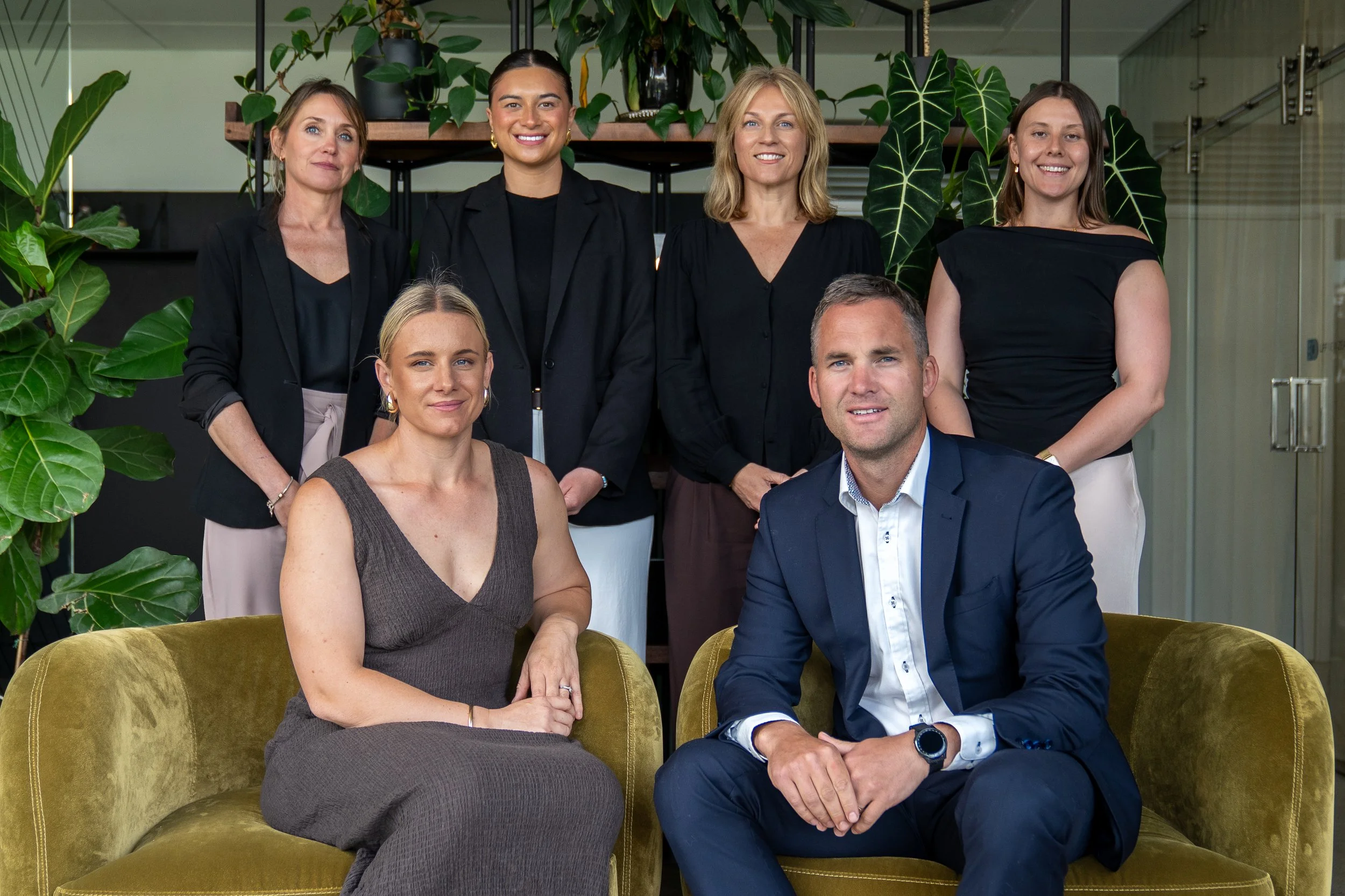 A group of seven professionally dressed men and women posing for a photo in an office with green plants in the background, sitting and standing around a mustard-colored velvet sofa.