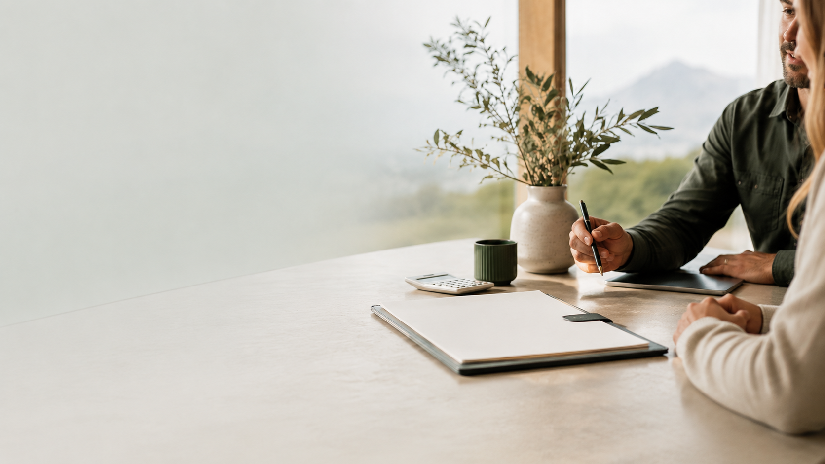 Two people engaged in a conversation at a desk, with a notebook, calculator, coffee cup, and a vase with green foliage, near a window showing a mountain view.