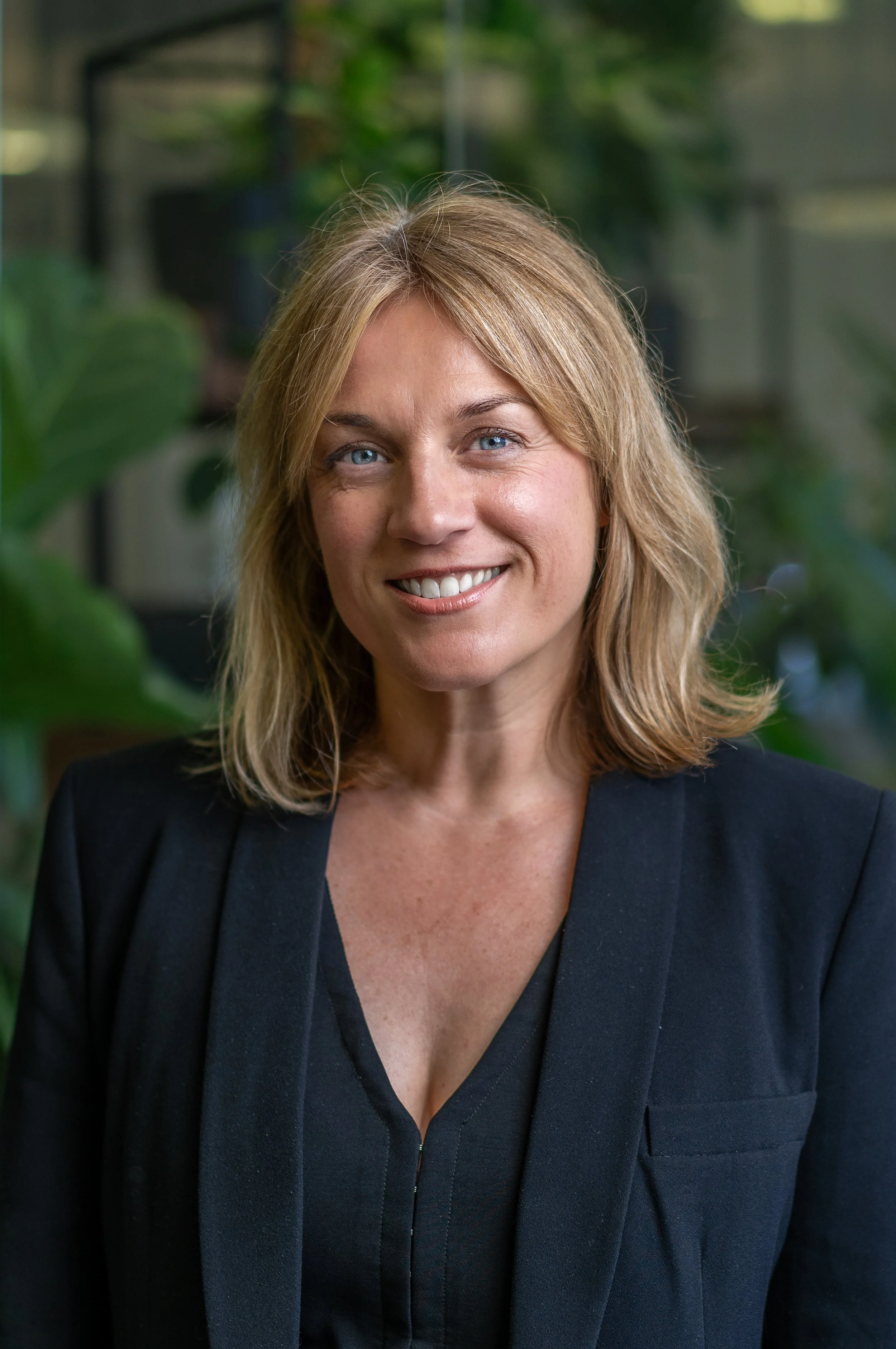A woman with blonde hair and blue eyes smiling, wearing a dark blazer, in an indoor setting with green plants in the background.