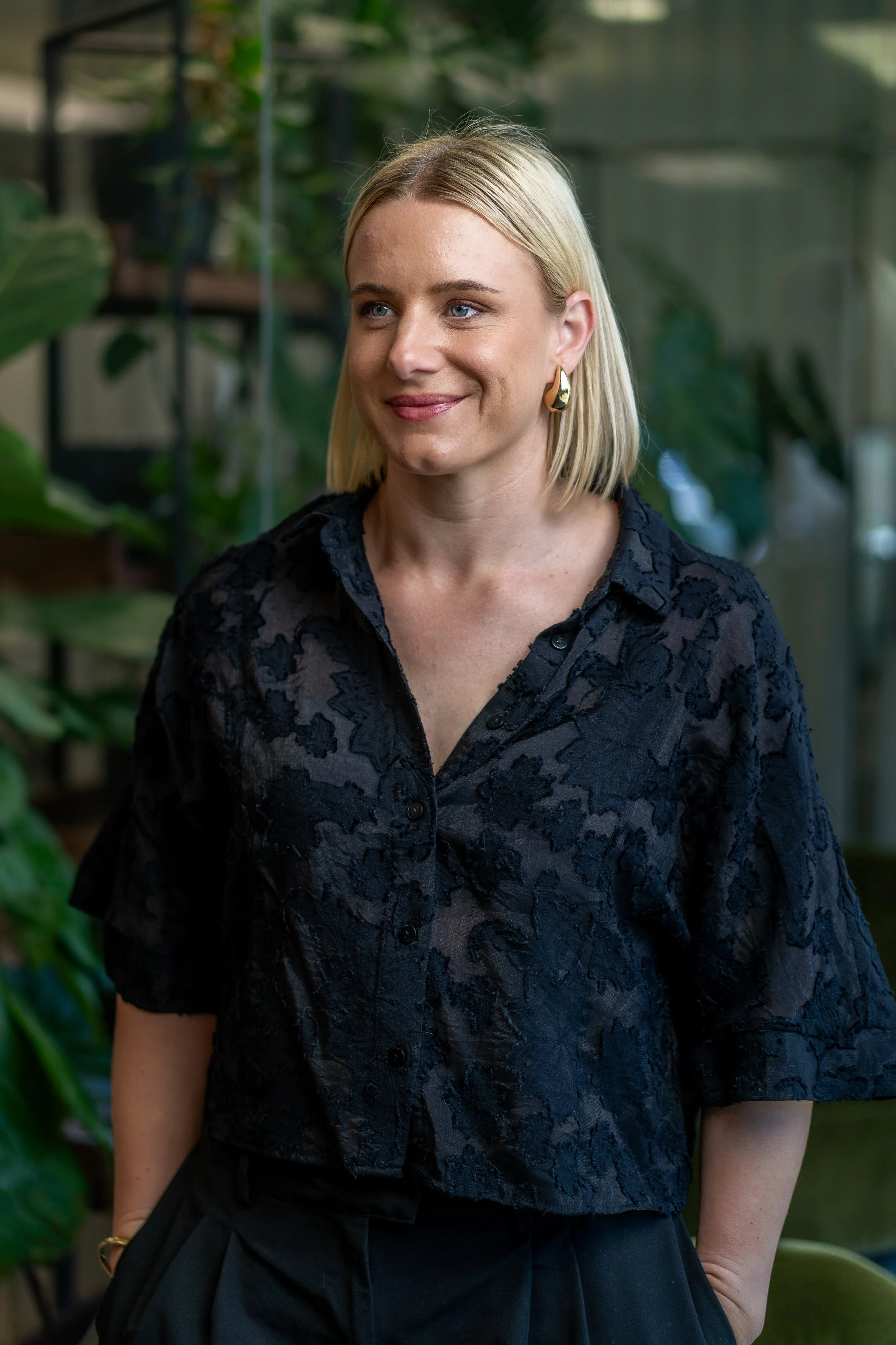 A woman with shoulder-length blonde hair, wearing a black lace top with a floral pattern and gold earrings, smiling and standing in front of green plants.