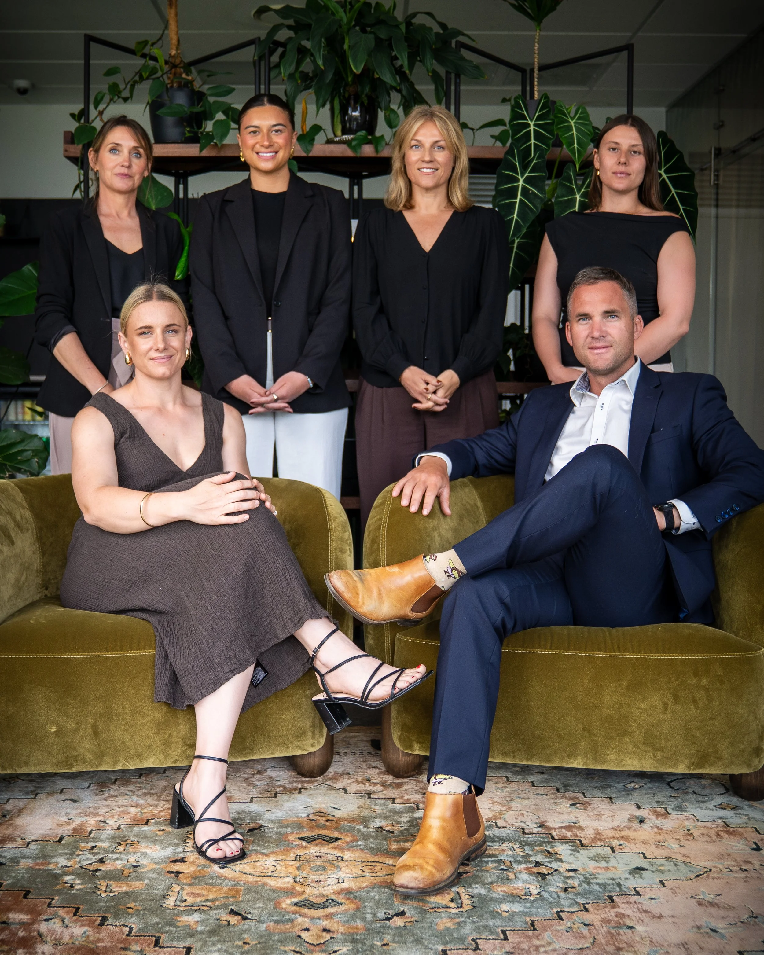 Five adults, four women and one man, posed inside a stylish indoor space with large green plants behind them. The women are dressed in black blazers and dresses, and the man is in a navy suit with a white shirt. They are seated and standing casually, smiling, with an olive-green velvet sofa and a patterned rug visible in the foreground.