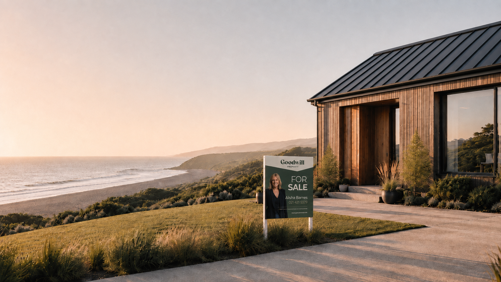 A modern house with wooden exterior and large windows located on a hillside overlooking the ocean during sunset. A 'SOLD' sign from Goodwill Property is in the front yard.