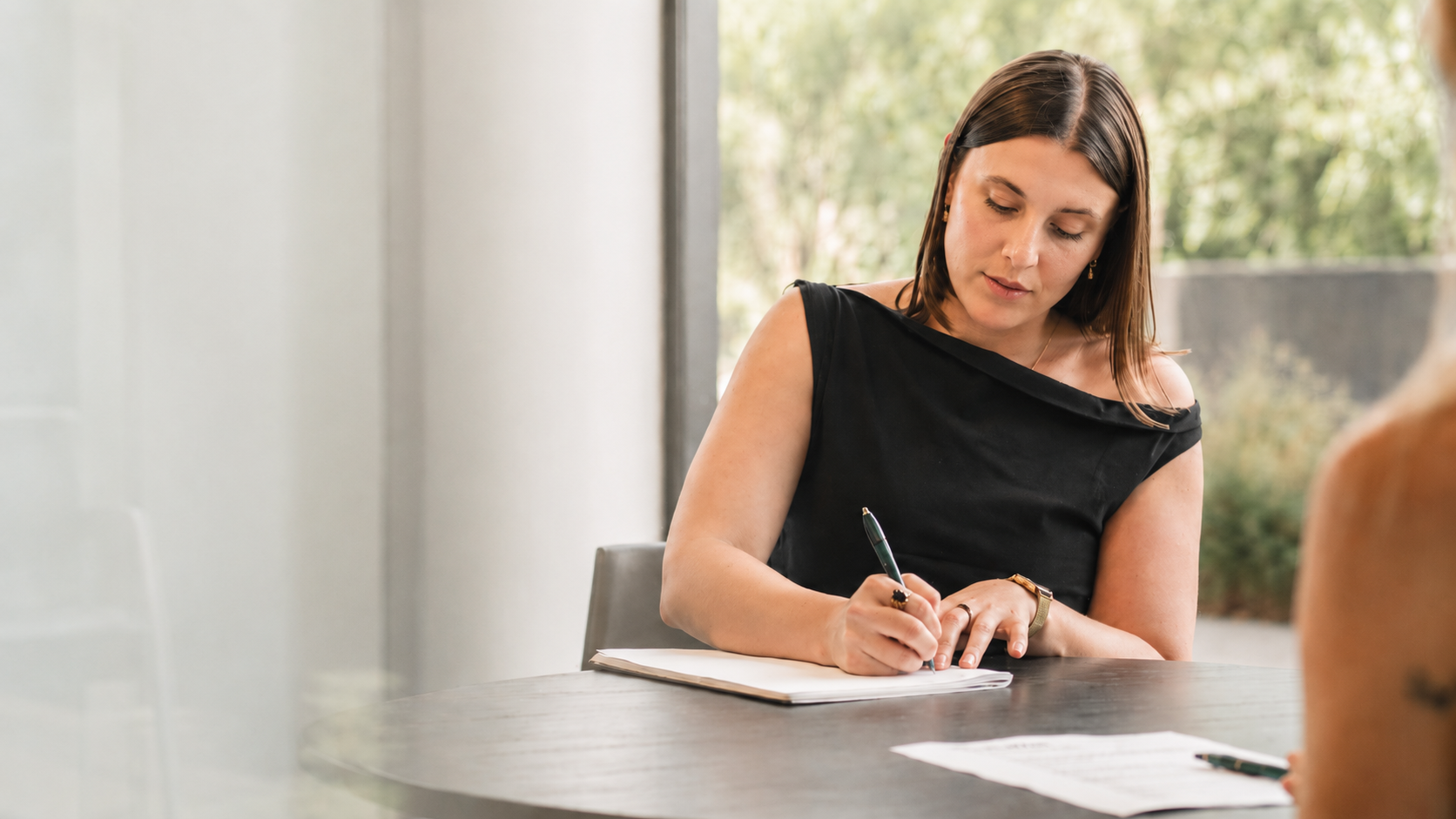 A woman with shoulder-length brown hair writing in a notebook during a meeting, sitting at a dark wooden table with some papers and a pen.