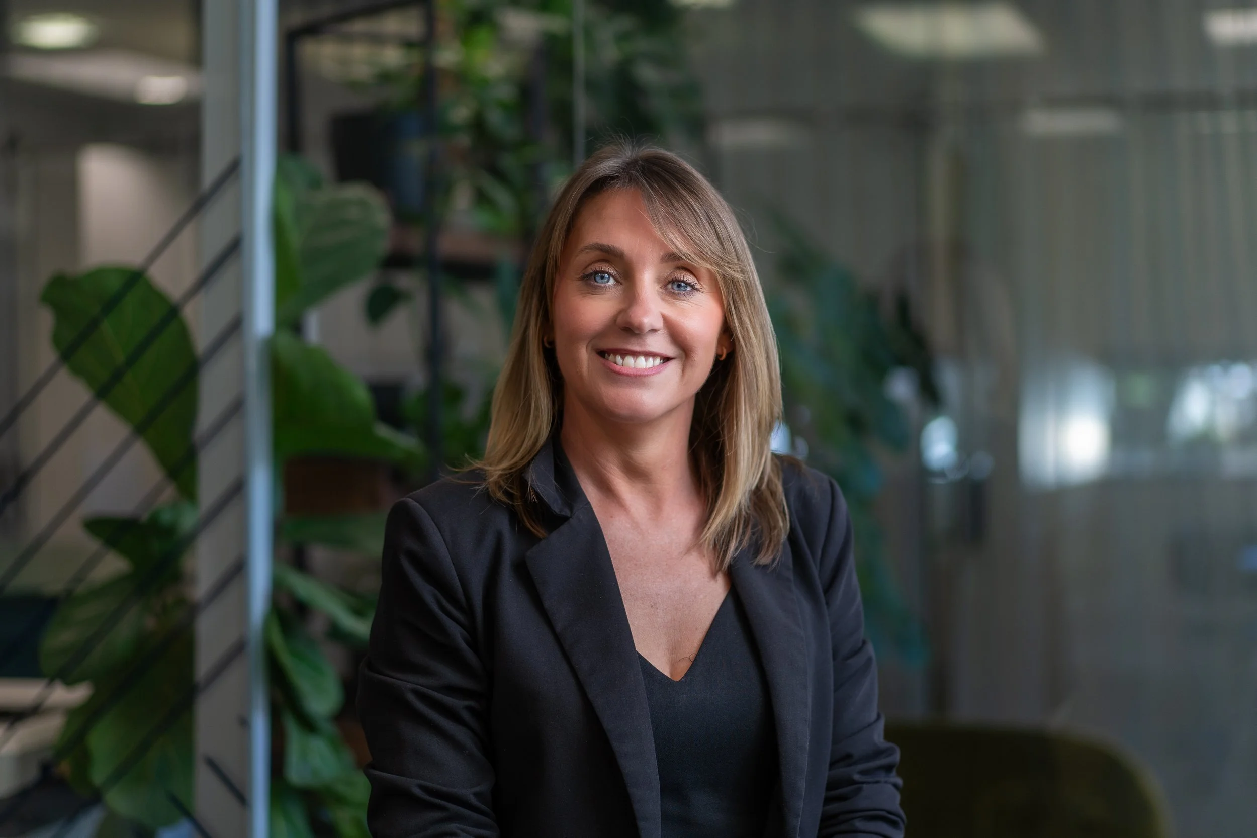A woman with shoulder-length blonde hair and blue eyes smiling, wearing a black blazer, standing in an indoor setting with green plants and a modern background.