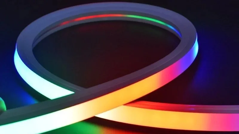 Close-up of a neon LED light strip displaying rainbow colors, arranged in a curved shape on a black surface.