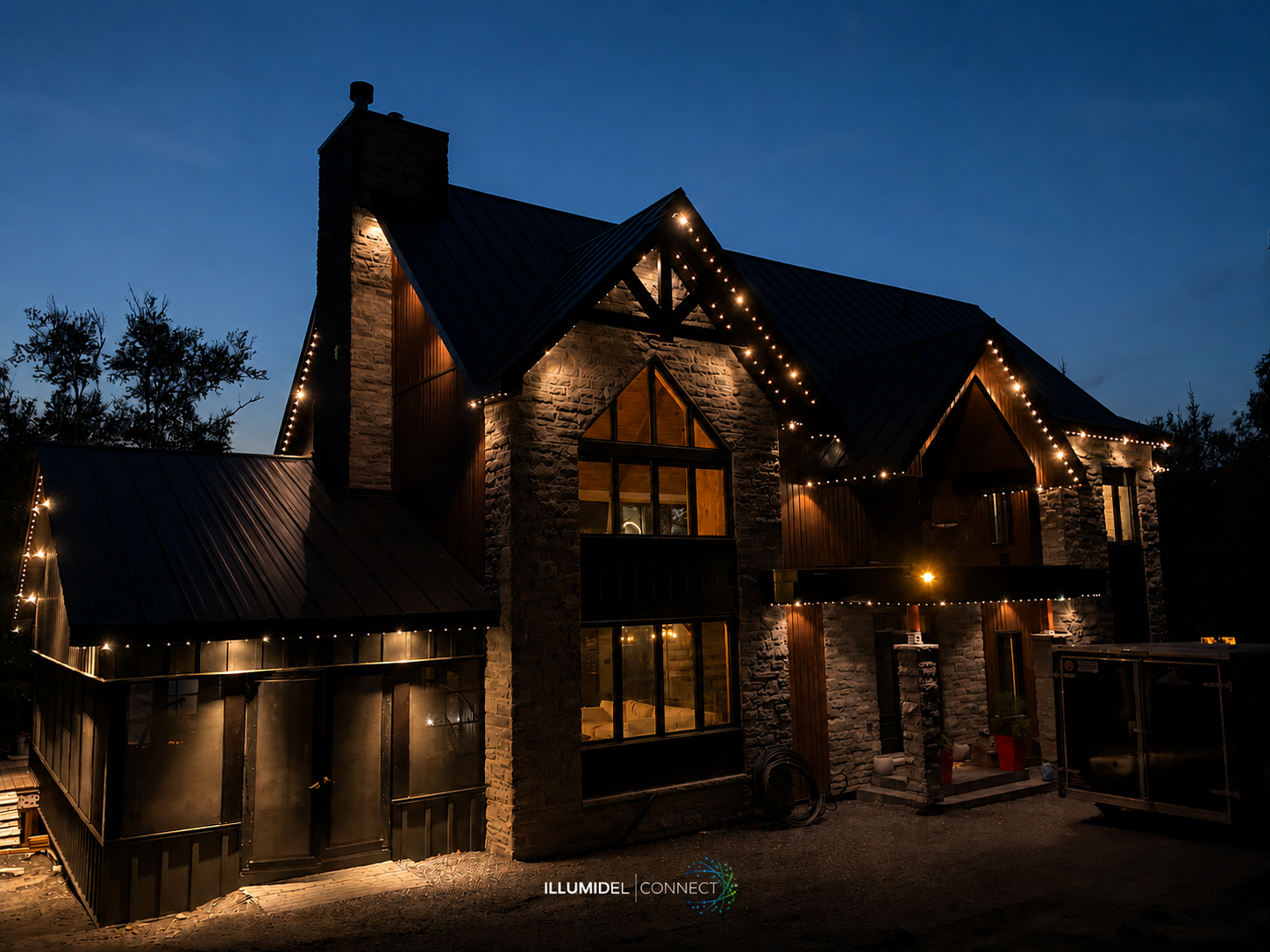 A large house at night decorated with string lights, featuring stone and wood siding, multiple windows, and a sloped metal roof.