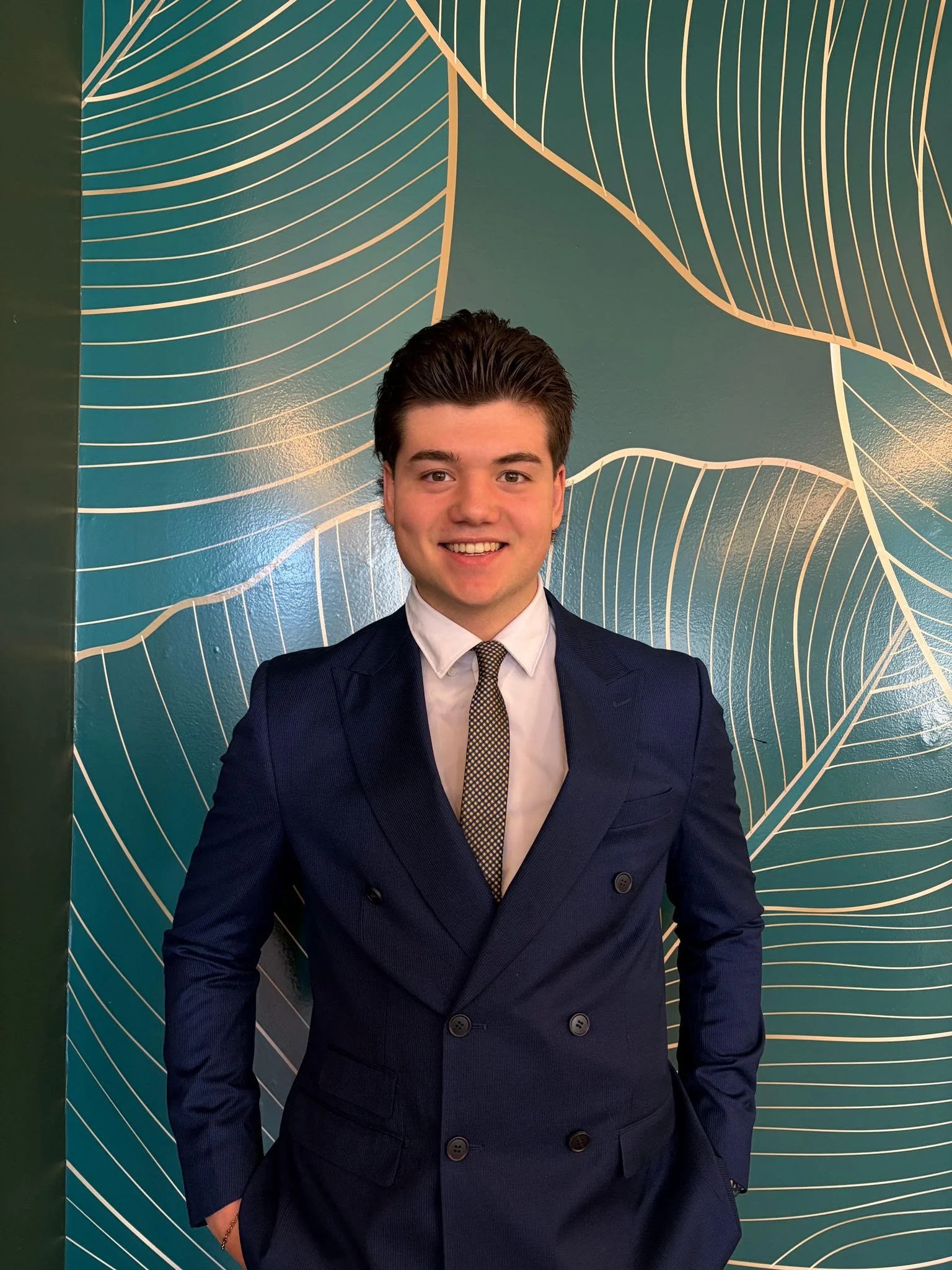 Young man in a dark blue business suit and patterned tie smiling, standing in front of a blue-green wall with white abstract line art design.