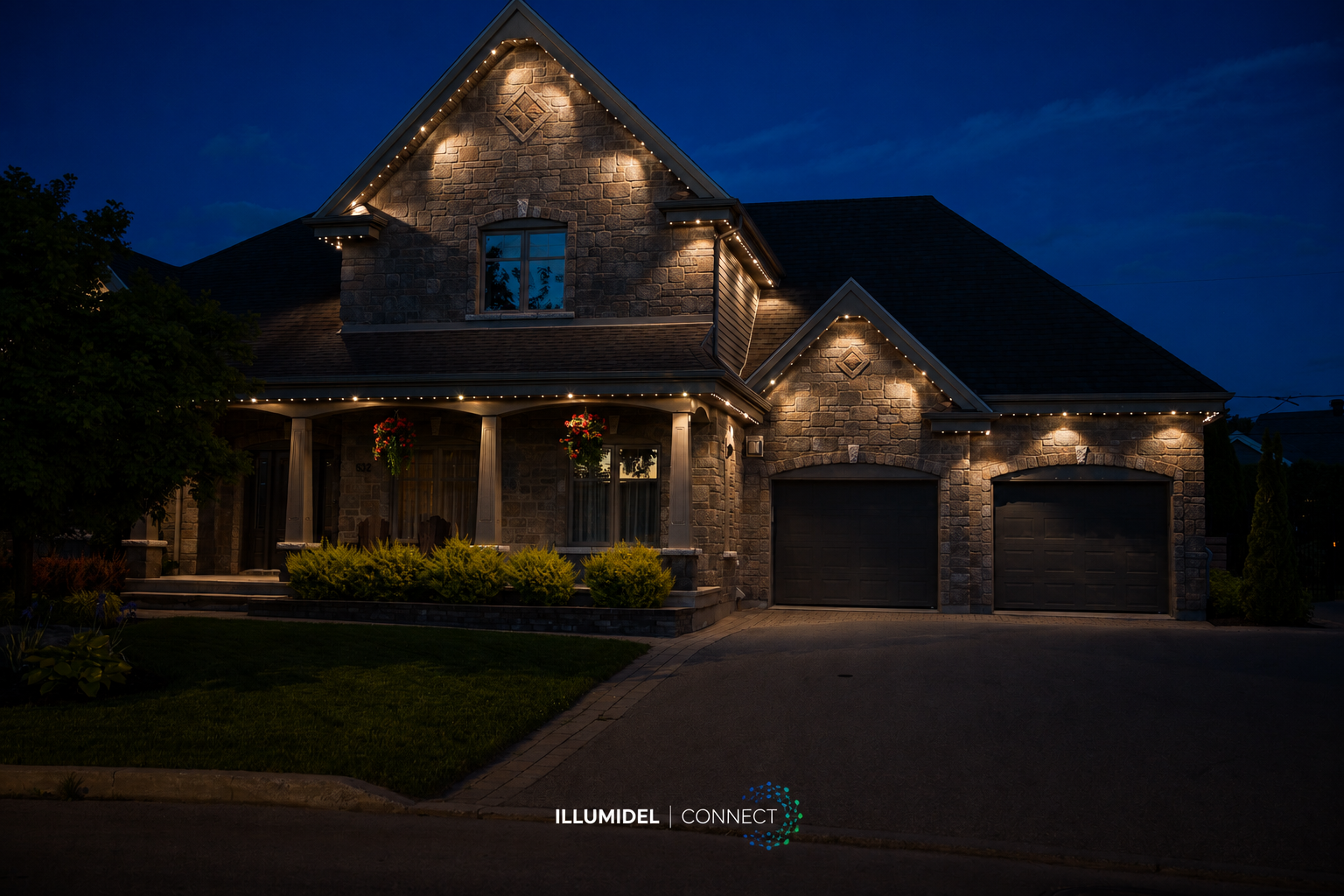 A large, two-story house illuminated with outdoor lighting during nighttime, featuring stone and siding exterior, front porch with hanging flower baskets, and a two-car garage.
