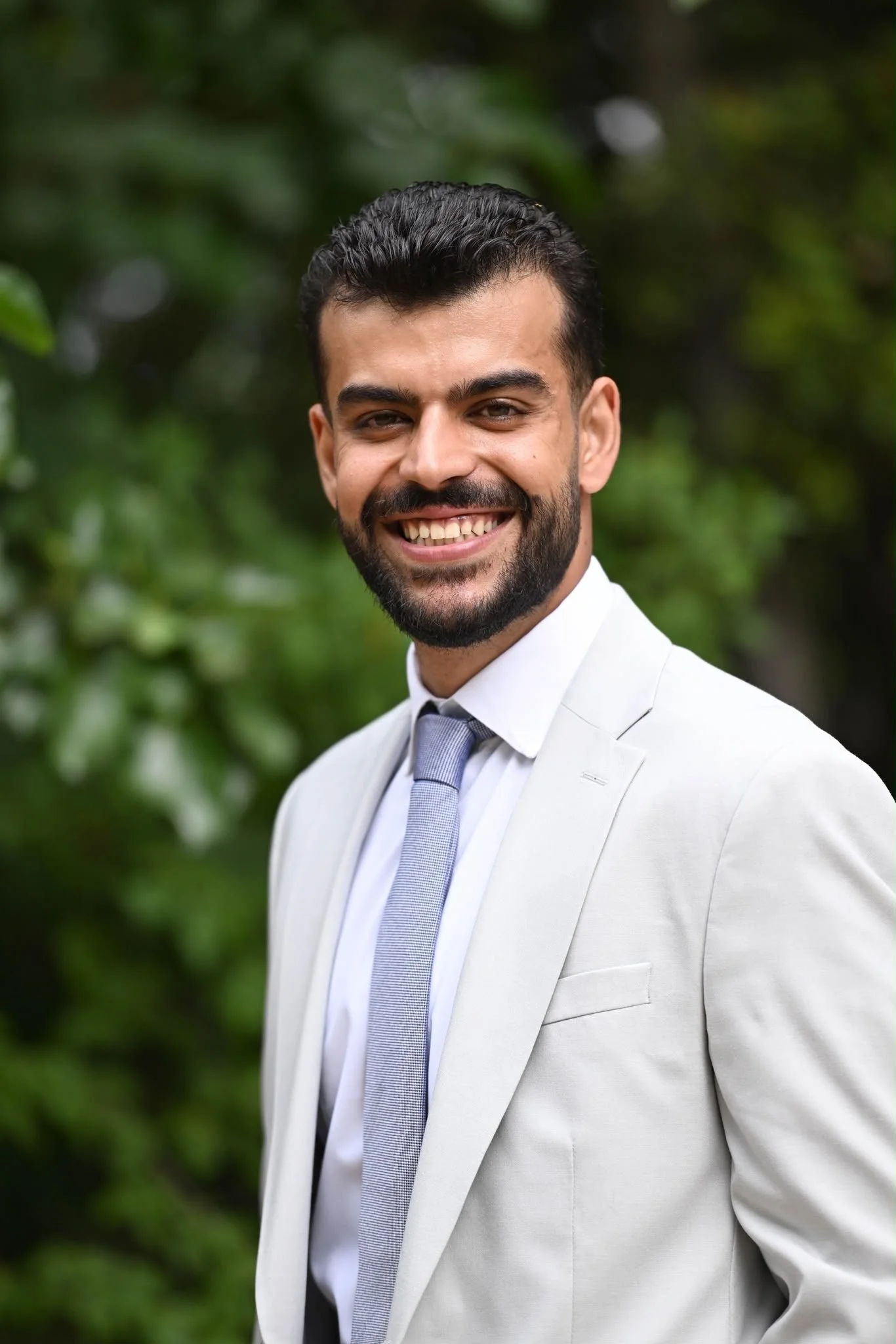 A smiling man in a light gray suit with a white shirt and gray tie, standing outdoors with blurred green foliage in the background.
