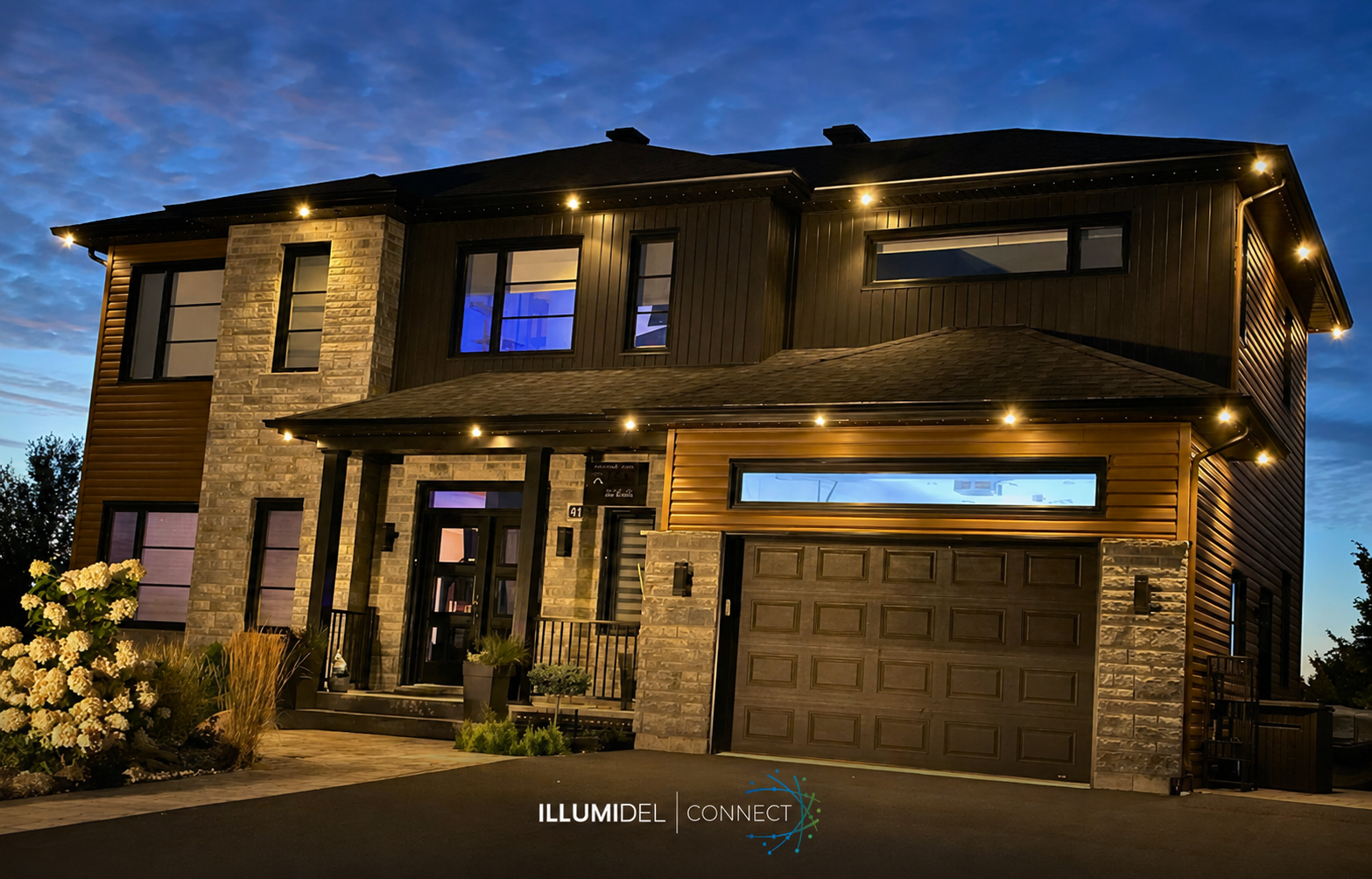 A modern two-story house illuminated at dusk with outdoor lighting, featuring brick and wood exterior, a garage, and landscaped front yard.