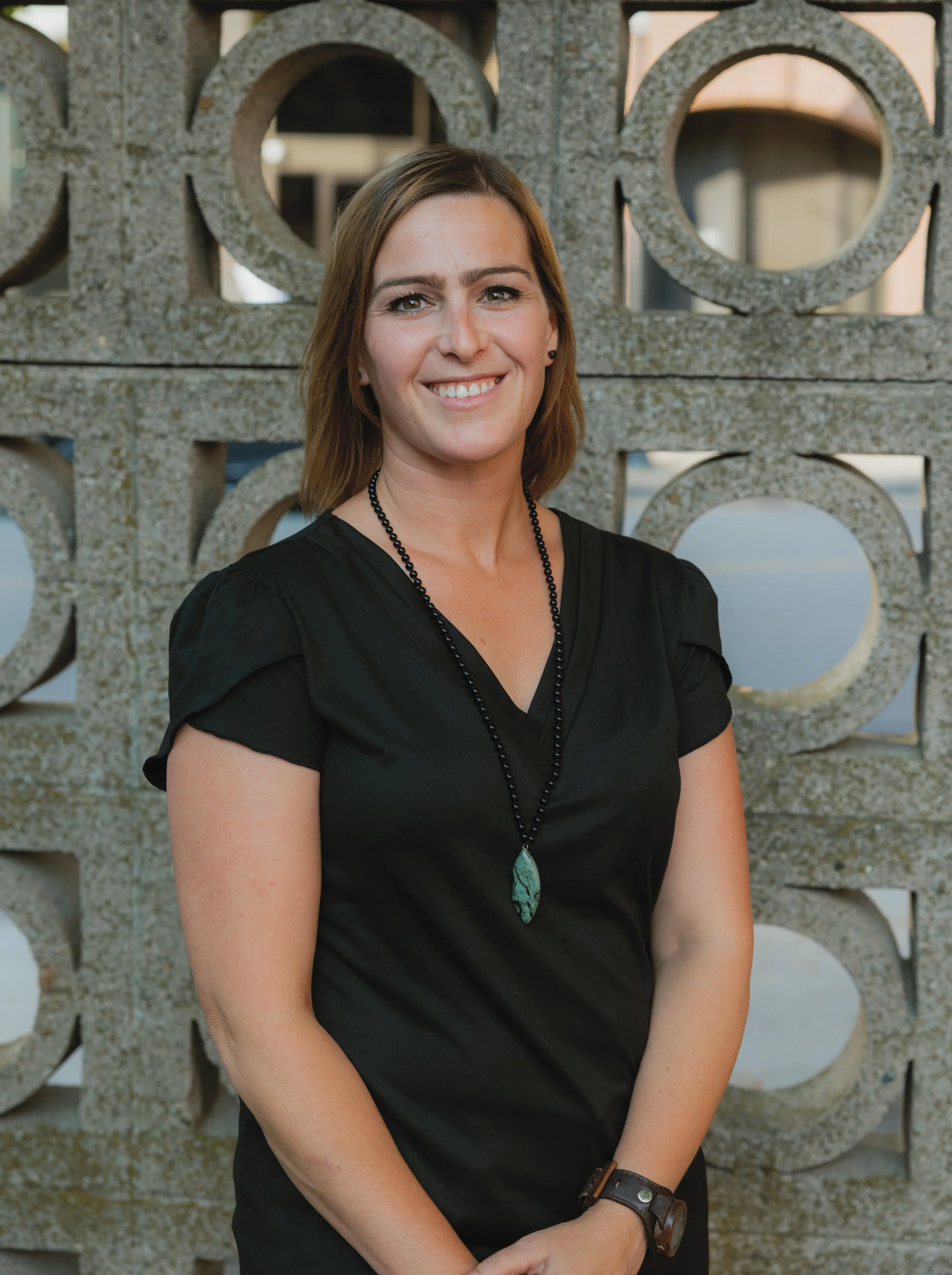 A woman with shoulder-length brown hair wearing a black top, necklace, and watch, standing in front of a decorative concrete wall with circular cutouts, smiling at the camera during daytime.