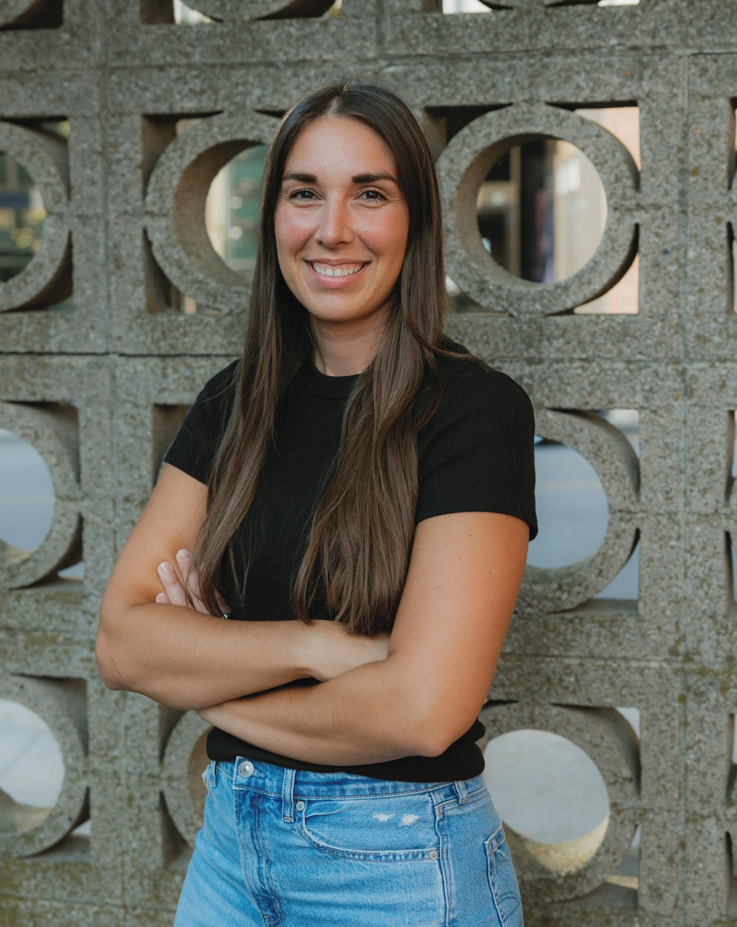 A young woman with long brown hair, wearing a black t-shirt and blue jeans, standing with arms crossed in front of a decorative concrete wall with circular cutouts.
