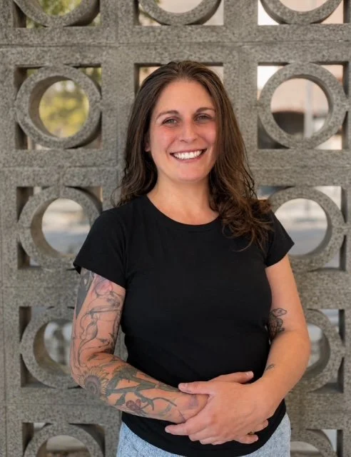 A woman with long brown hair, tattoos on her arms, smiling and wearing a black t-shirt, standing in front of a decorative concrete wall with circular cutouts.