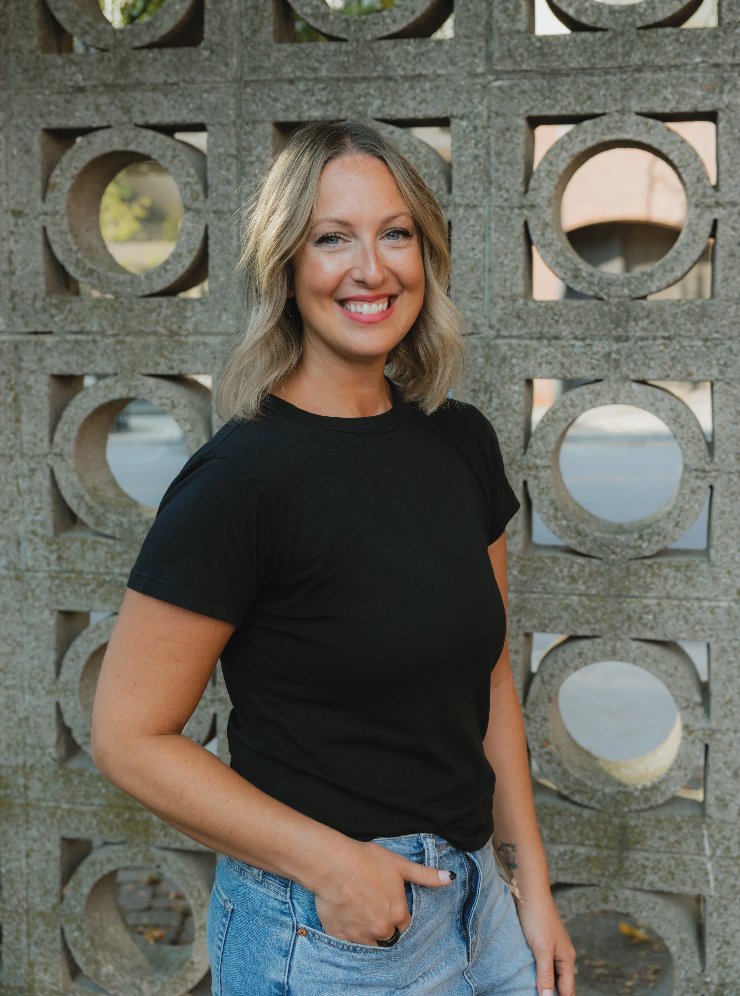 A woman with blonde hair, blue eyes, and a bright smile standing outdoors by a decorative concrete wall with circular cutouts. She is wearing a black T-shirt and blue jeans, and has a small tattoo on her left wrist.