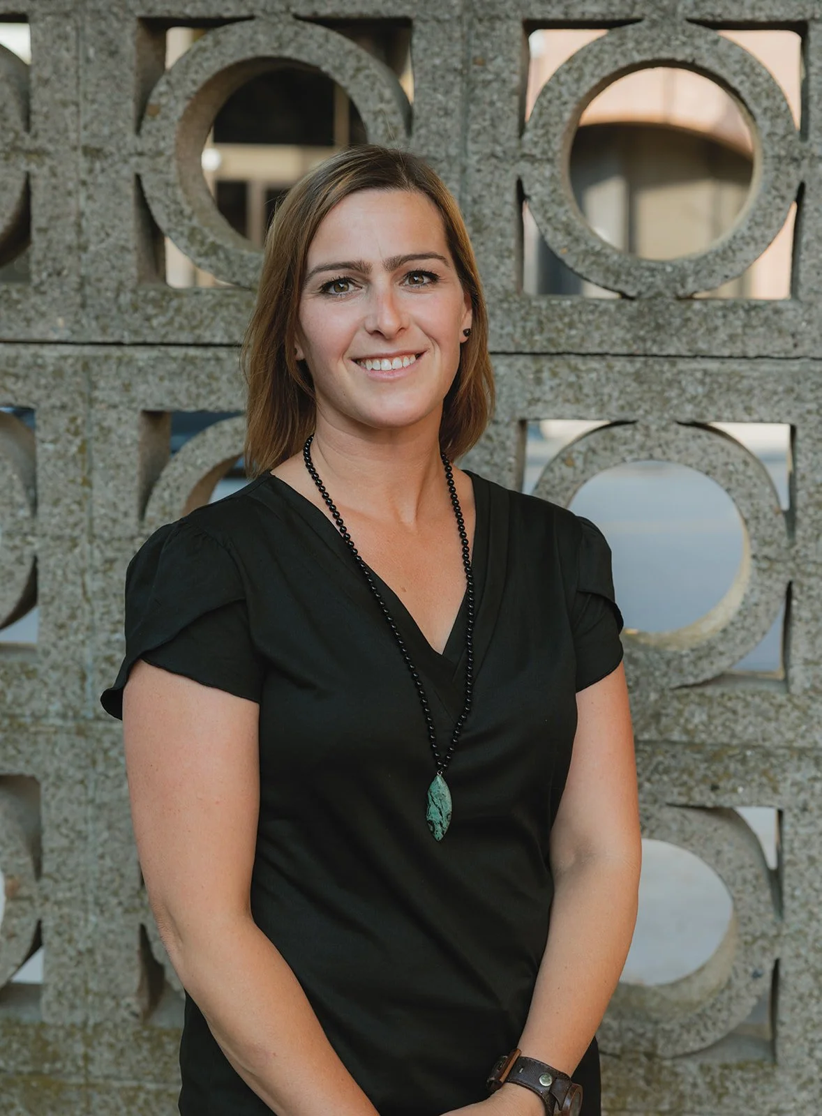 A woman with shoulder-length brown hair, wearing a black top and a black beaded necklace with a turquoise pendant, standing in front of a decorative concrete wall with circular cutouts.