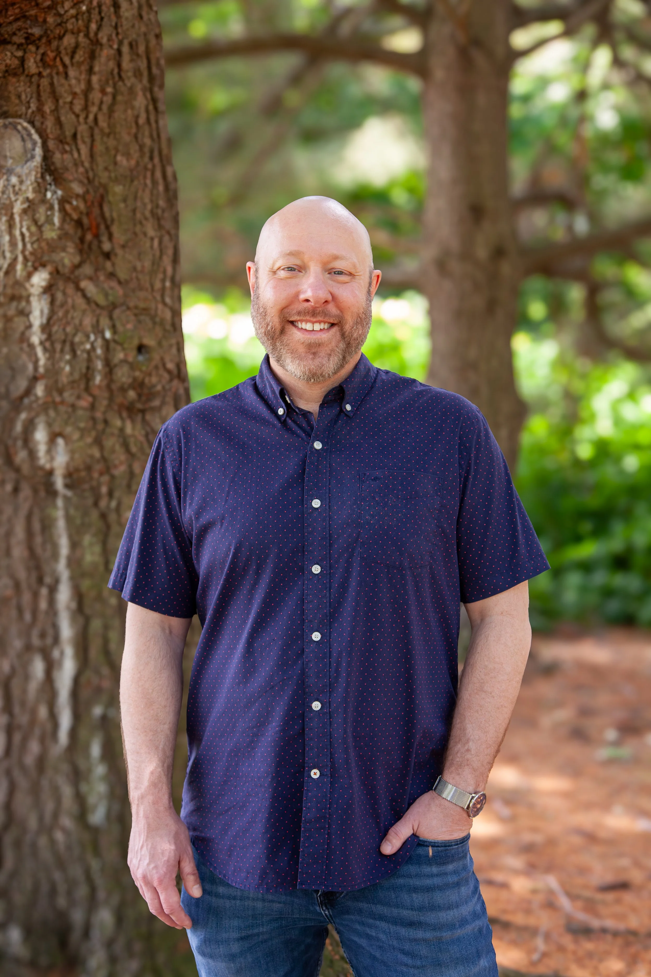 A smiling man with a beard and bald head wearing a navy blue short-sleeved button-up shirt with small red dots, standing outdoors in front of a large tree with a blurred forest background.